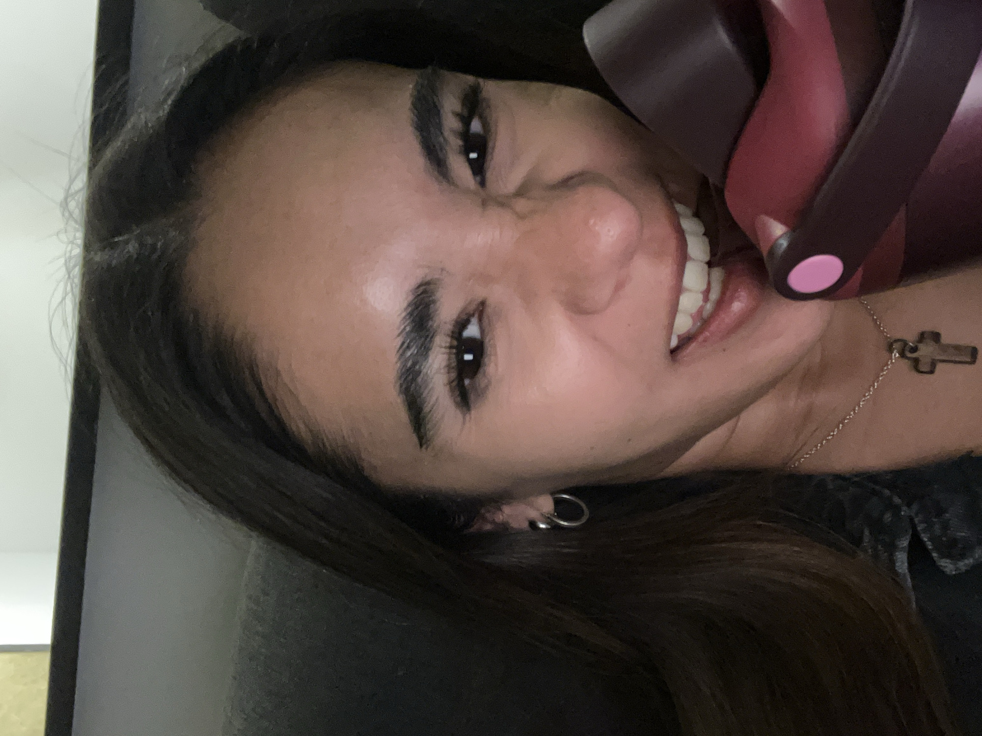 A person with dark hair and brown eyes is close to the camera, showing teeth in a grimace. A pink and dark maroon-colored object is close to their face. The person is also wearing a silver cross necklace and a silver earring. The setting appears to be indoors. The city of San Bernardino Tlaxcalancingo, Mexico, is not visibly indicated in the image.