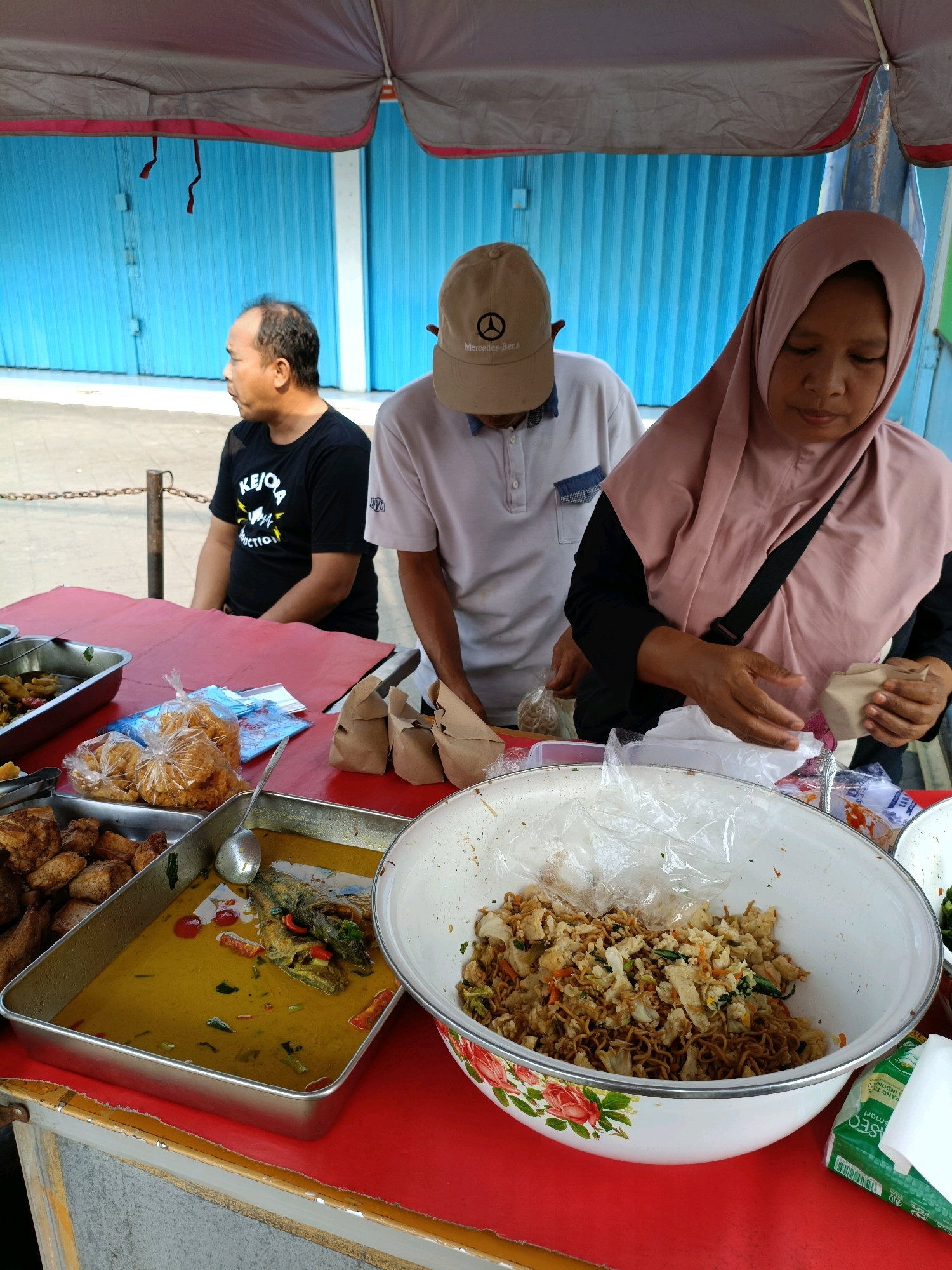 In Maguwoharjo, Indonesia, three individuals are positioned at a food stall with a red table beneath a gray and pink striped awning. On the left, a bald man with dark clothing sits looking to the left. In the center, a person wearing a beige cap with a Mercedes-Benz logo and a light-colored polo shirt stands, appearing to serve food. On the right, a woman wearing a pink hijab and dark clothing handles a small brown paper-wrapped package. The table displays various food items in metal trays and bowls. One tray contains fried items, another holds fish in a yellow curry, and a large bowl is filled with stir-fried noodles and vegetables. Several brown paper bags and plastic-wrapped snacks are also visible. The background features a bright blue corrugated metal wall and a chain link fence.