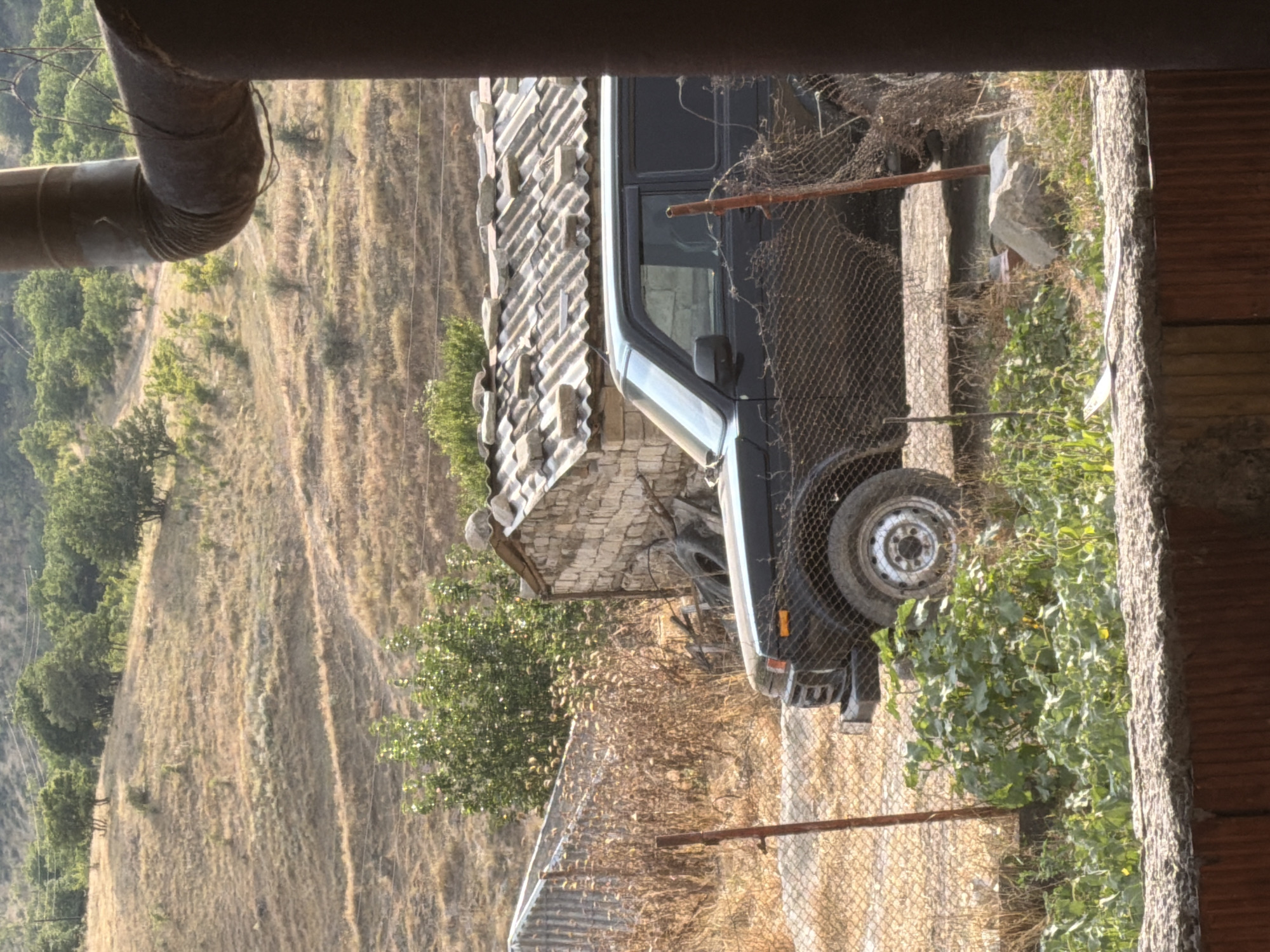 The image depicts a rural outdoor scene in Köhnə Quşçu, Azerbaijan, dominated by a black off-road vehicle parked behind a wire fence. The vehicle, likely a Lada Niva, is positioned in front of a small, rustic building with a corrugated roof covered in stones, and a wall constructed of rough-hewn stones. In the foreground, lush green vegetation grows. The background features a dry, hilly landscape with sparse trees and vegetation, indicative of a warm, arid climate. A large, rusted metal pipe is prominently visible in the right side of the frame, partially obscuring the view of the landscape and the sky. There are no people visible in the image.