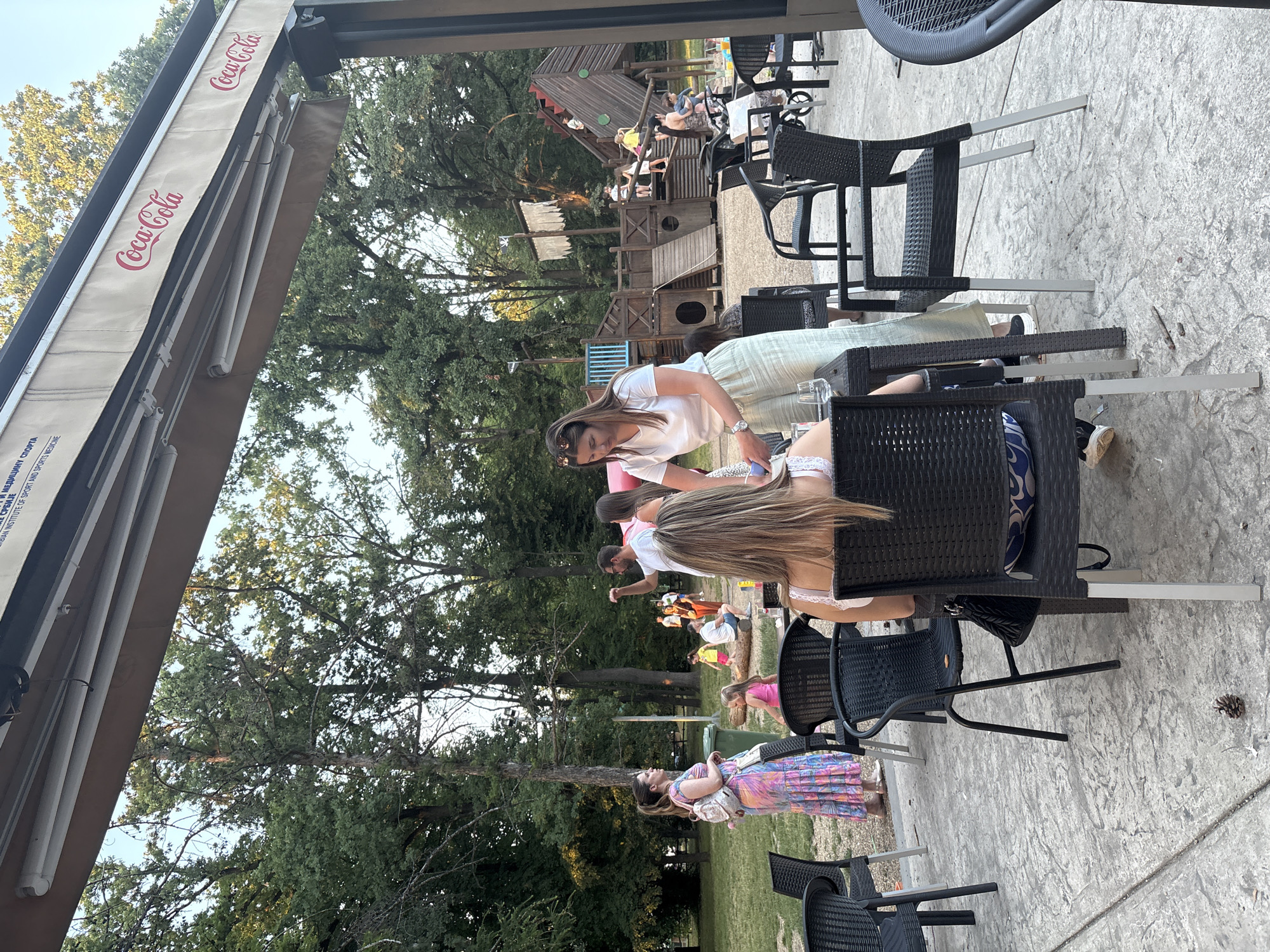 The image depicts an outdoor seating area at a cafe or restaurant with a children's playground in the background. The cafe has black wicker chairs with silver legs, and tables with stone-patterned tops. An awning with Coca-Cola branding is visible overhead, suggesting the cafe is located in the Cukarica Urban Municipality, Serbia, based on the information provided in the query.
In the foreground, two women are seated at a table, facing away from the camera. One woman has blonde hair and is wearing a white top with a blue and white patterned cushion on her chair. The other woman, closer to the camera, is partially visible from the back, wearing a white top. Another woman, standing and facing left, is wearing a colorful, long, patterned dress and a light-colored backpack. She appears to be interacting with someone off-camera.
The background features a large wooden playground designed like a pirate ship, with slides, ladders, and platforms. Several adults and children are visible in and around the playground, indicating a family-friendly environment. Trees and greenery surround the area, suggesting an outdoor park or recreational space. The ground is paved with a textured stone material. The overall scene suggests a casual, public gathering place, likely a cafe or restaurant adjacent to a park or playground.