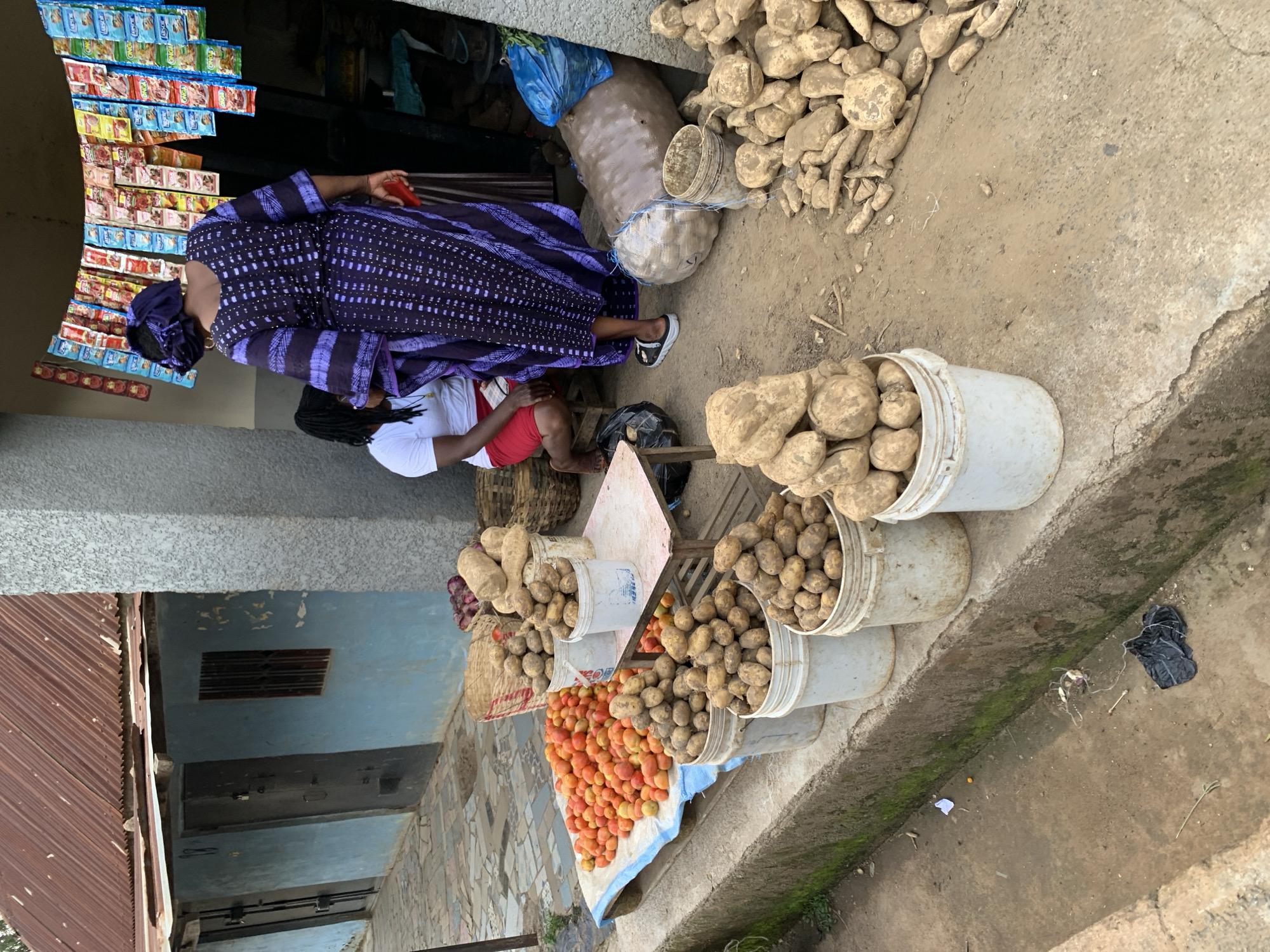 In an outdoor market scene in Jos, Nigeria, a woman wearing a traditional purple and black patterned dress and headscarf is seen standing. She is holding a red object in her right hand, possibly a piece of fruit or a small item. To her left, a person in a white t-shirt and red shorts is seated on a wooden structure, with their back to the camera.
The market stall features an array of goods. In the background, shelves are stacked with colorful packaged items, likely snacks or small food products, with visible text that is illegible. In the foreground, a large pile of yams sits on the ground, and several white buckets are filled with potatoes. A blue tarp is spread out on the ground, laden with a vibrant mound of red tomatoes. Various baskets and bags are also visible, holding more produce.
The ground appears to be a mixture of dirt and concrete. A narrow, moss-lined trench runs along the right side of the image, with a crumpled black plastic bag lying near it. The scene suggests a bustling market day, with produce laid out for sale. The lighting indicates it is daytime, though specific time or weather conditions are not evident. The overall atmosphere appears to be one of commerce and daily life in a Nigerian market.