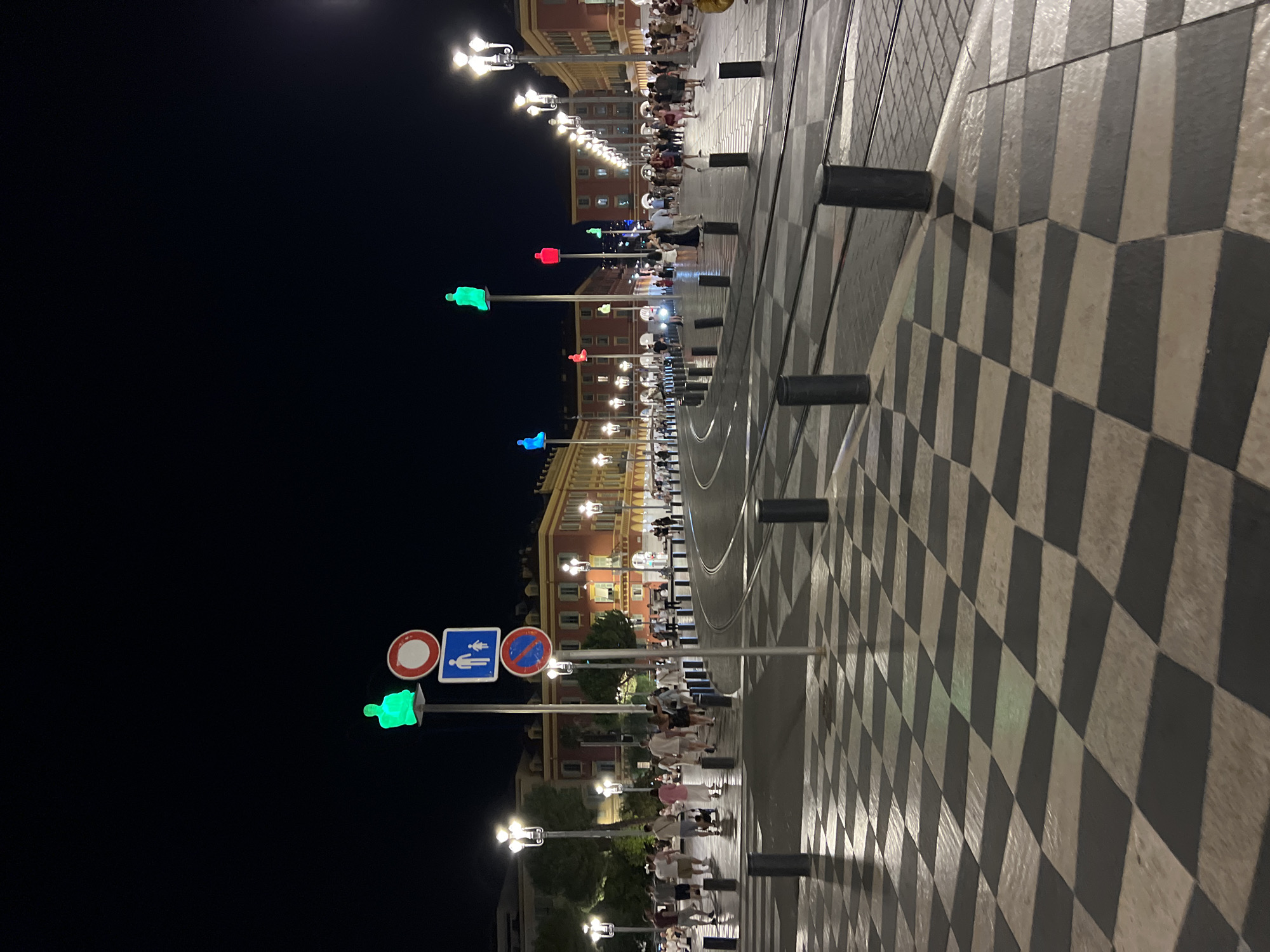 This image captures a nighttime scene on the Promenade du Paillon in Nice, France. The foreground is dominated by a striking black and white checkered pavement pattern that reflects the surrounding lights. Several dark cylindrical bollards are placed along the edge of the pedestrian walkway. In the middle ground, a streetcar track curves through the plaza.
A prominent traffic signal pole stands in the left-center of the frame, displaying a green pedestrian silhouette, a circular red and white prohibition sign, and a blue square sign with a white pedestrian icon. Further into the plaza, a series of tall poles are topped with illuminated human figures in various colors, including green, blue, and red, creating a vibrant artistic display against the dark night sky.
Numerous people are visible walking and standing on the promenade, dressed in casual attire. They are illuminated by the streetlights and the colored figures. The background features elegant, multi-story buildings with red facades and numerous windows, typical of Nice's architecture. The atmosphere is lively, suggesting a popular public space in the evening. The lighting creates reflections on the wet pavement, adding to the dynamic visual quality of the scene.