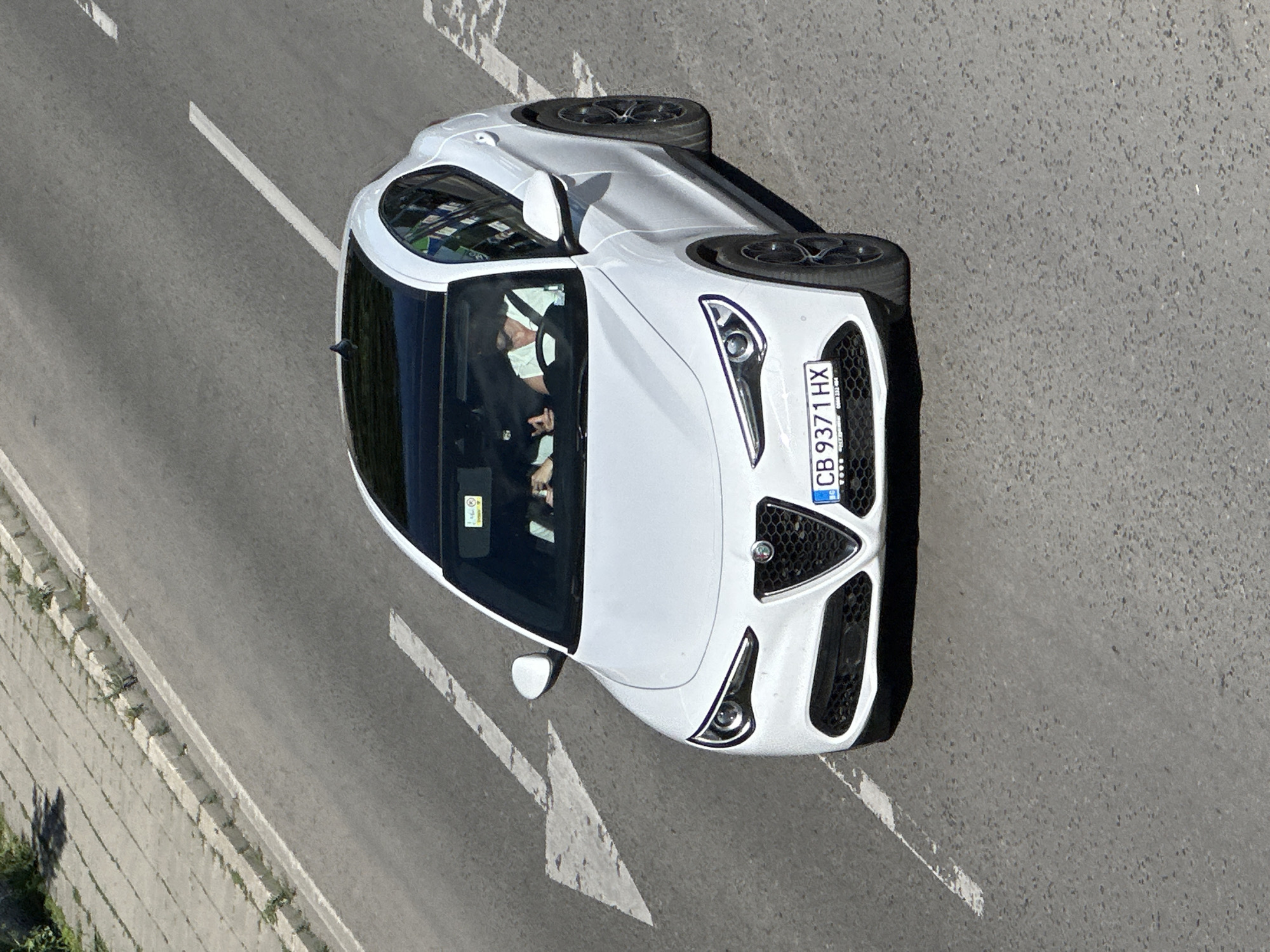 A white Alfa Romeo Stelvio is driving on a paved road in Veliko Tarnovo, Bulgaria. The car has a black panoramic sunroof and tinted windows. Two people are visible inside the car, the driver and a passenger in the front seat. The driver is holding the steering wheel. The license plate of the car reads "CB 9371 HX". The road appears to be a multi-lane street with white lane markings. There is a sidewalk and some vegetation on the left side of the road. The sky is clear and the lighting suggests it is daytime.