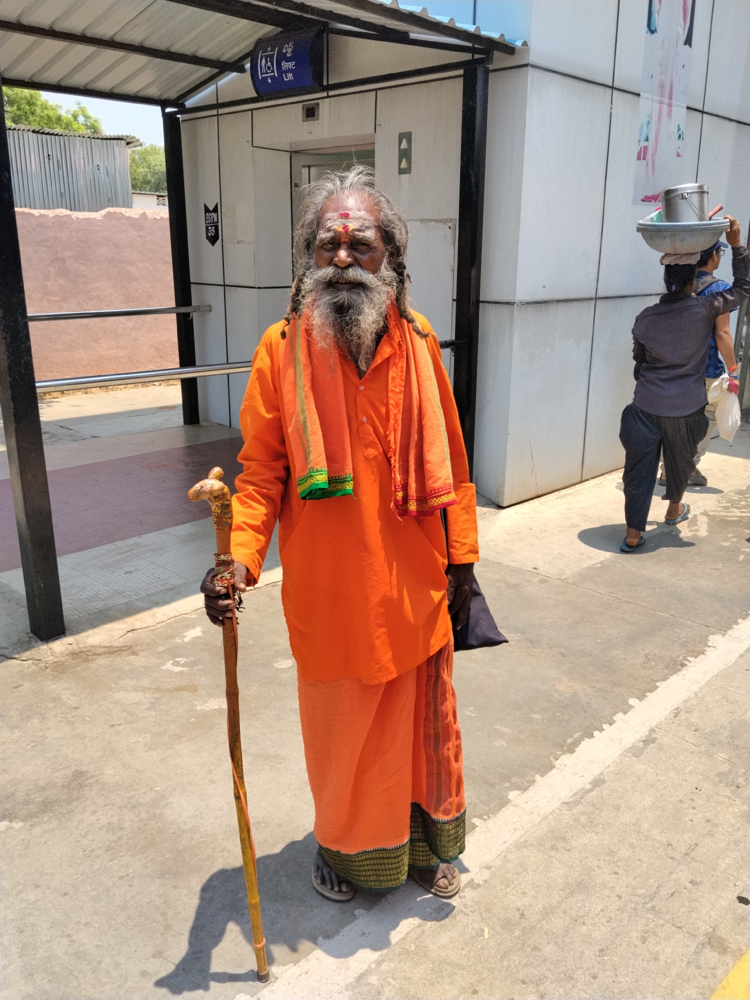 The image shows an Indian man in an orange robe standing in front of a building. He has a long beard and is holding a wooden staff. Behind him, a person is carrying items on their head. The setting appears to be in Mamillakunta, India. There are signs with writing on the building.