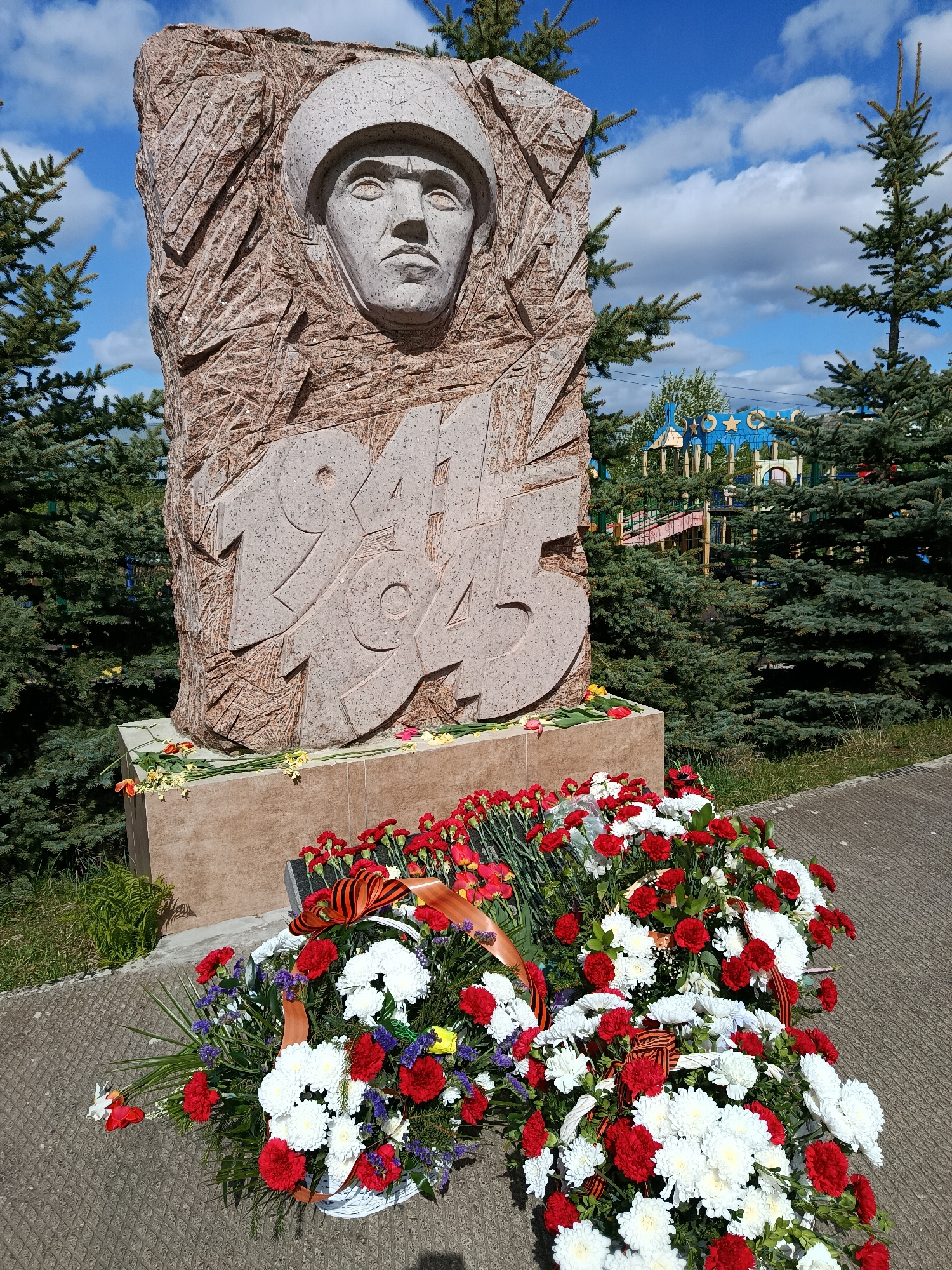 The image depicts a stone monument dedicated to the year 1945, likely commemorating the end of World War II. Carved into the stone is a soldier's face wearing a helmet. The base of the monument is adorned with numerous red and white carnations and other flowers, arranged in wreaths and bouquets, as well as St. George's ribbon. The monument is situated in an outdoor setting, likely a park or memorial site, with evergreen trees and other greenery visible in the background. There is a blue and yellow structure that appears to be a playground behind the trees. The location is the city of Фоки, Russia. The day is sunny with some clouds.