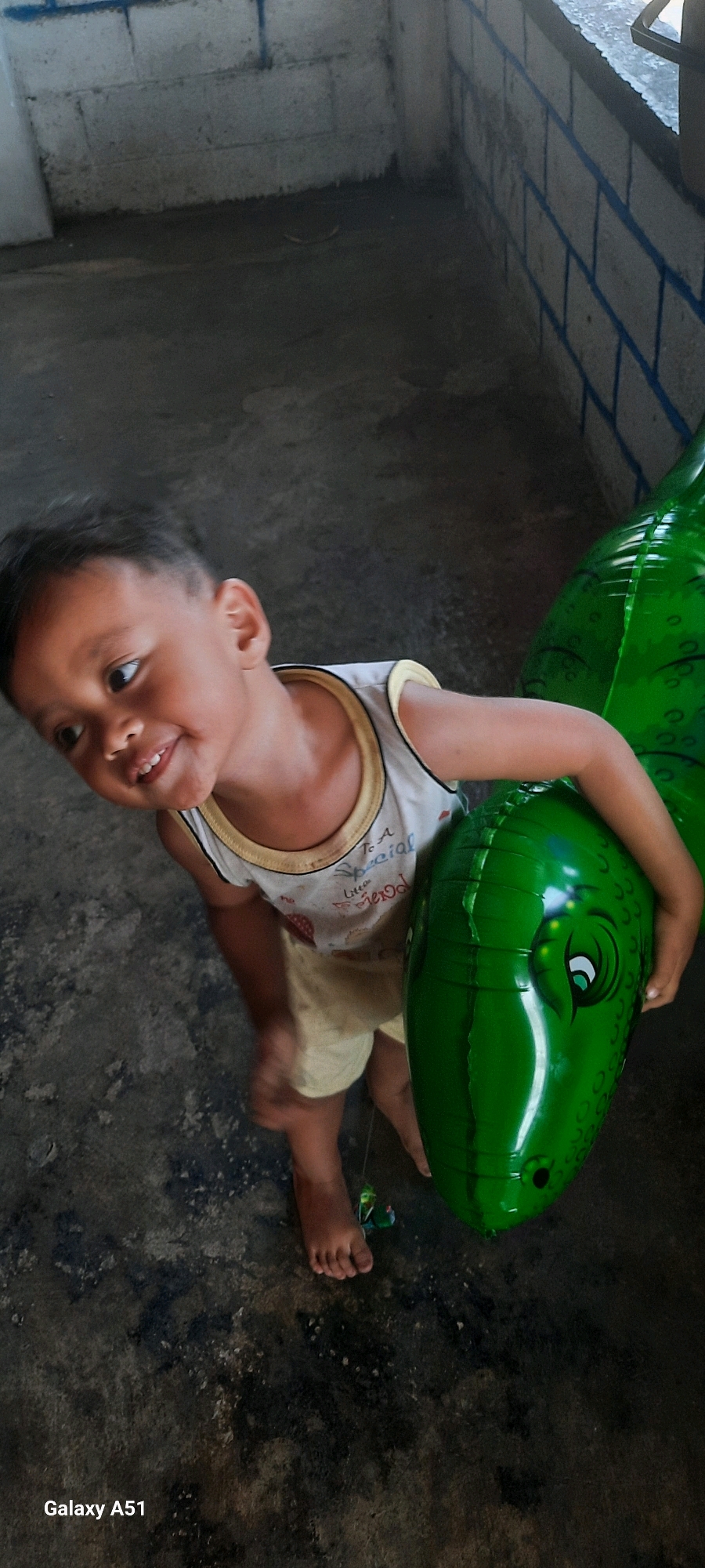 In the city of Selopuro, Indonesia, a young child is pictured outdoors, likely on a concrete surface, holding a large, green, inflatable dinosaur toy. The child, with dark hair and wearing a white tank top with a yellow trim and light-colored shorts, is smiling and looking to their left. The dinosaur toy's head, featuring a cartoonish eye and mouth, is prominently displayed. The background shows a textured concrete floor and a wall constructed of cinder blocks, some of which are painted with blue stripes. The lighting suggests it is daytime.