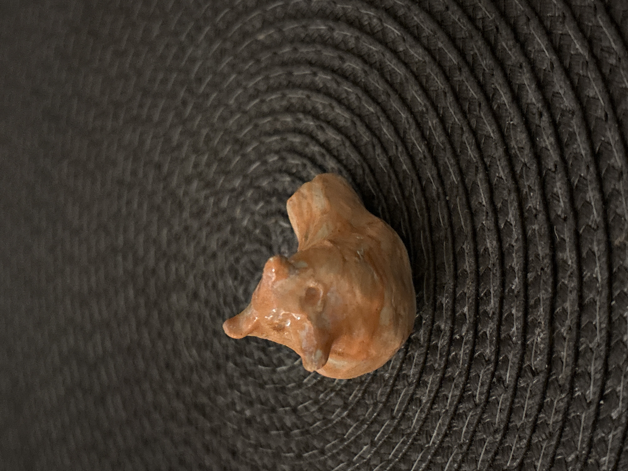 A small, ceramic animal figurine rests on a gray, spiral-woven mat. The figurine is light brown, rounded, and appears to depict a sleeping animal, possibly a pig or bear. The location is unknown and there is no evidence to suggest the city is Saint Petersburg, Russia.