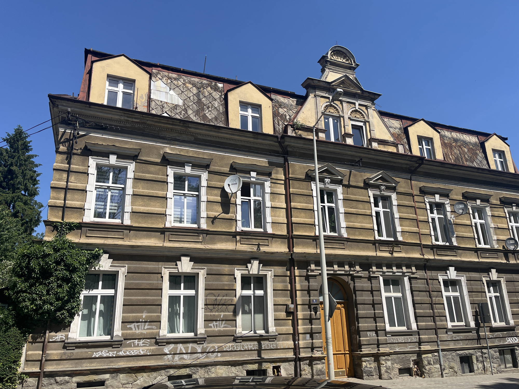 An eye-level, medium shot captures the facade of a weathered, historic building under a clear blue sky. The building is a multi-story structure with a steeply pitched, tiled roof. Dormer windows with yellow trim jut out from the roofline, some with white-framed panes. The building's exterior walls are painted a mottled brown, with horizontal banding and decorative white trim around the rectangular windows, which are predominantly white-framed. The lower portion of the facade transitions to a rough-hewn stone foundation. Graffiti is visible on the lower sections of the brown walls and on the stone foundation. A tall, thin lamppost stands in front of the building, positioned slightly off-center to the right. To the left of the building, a dense, green tree with ivy climbing its trunk partially obscures the facade. A yellow wooden door with a curved top is visible on the right side of the building, recessed into an archway. Several satellite dishes are attached to the upper sections of the facade. The street in front of the building is paved with grey material.