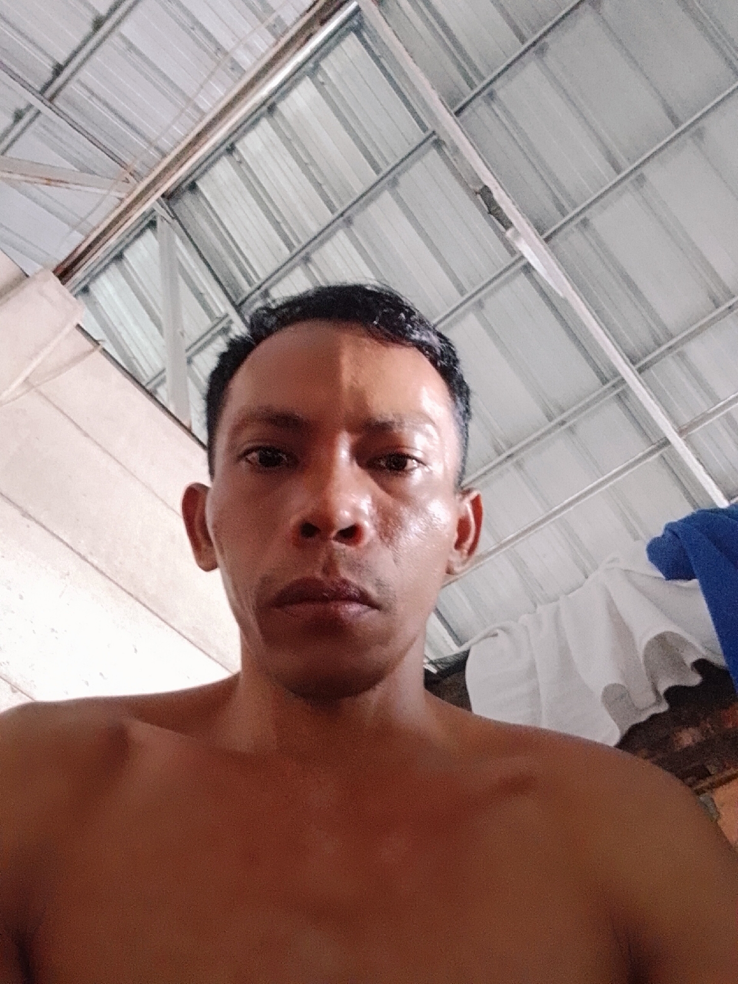 A medium close-up captures a man with dark, short hair and brown skin. He appears shirtless, and the photograph is taken from a low angle, looking up at him. Behind him is a ceiling made of corrugated metal, and a white and blue cloth. The location is possibly in the city of Bukit Ketok, Indonesia, given user metadata. The scene appears to be indoors.