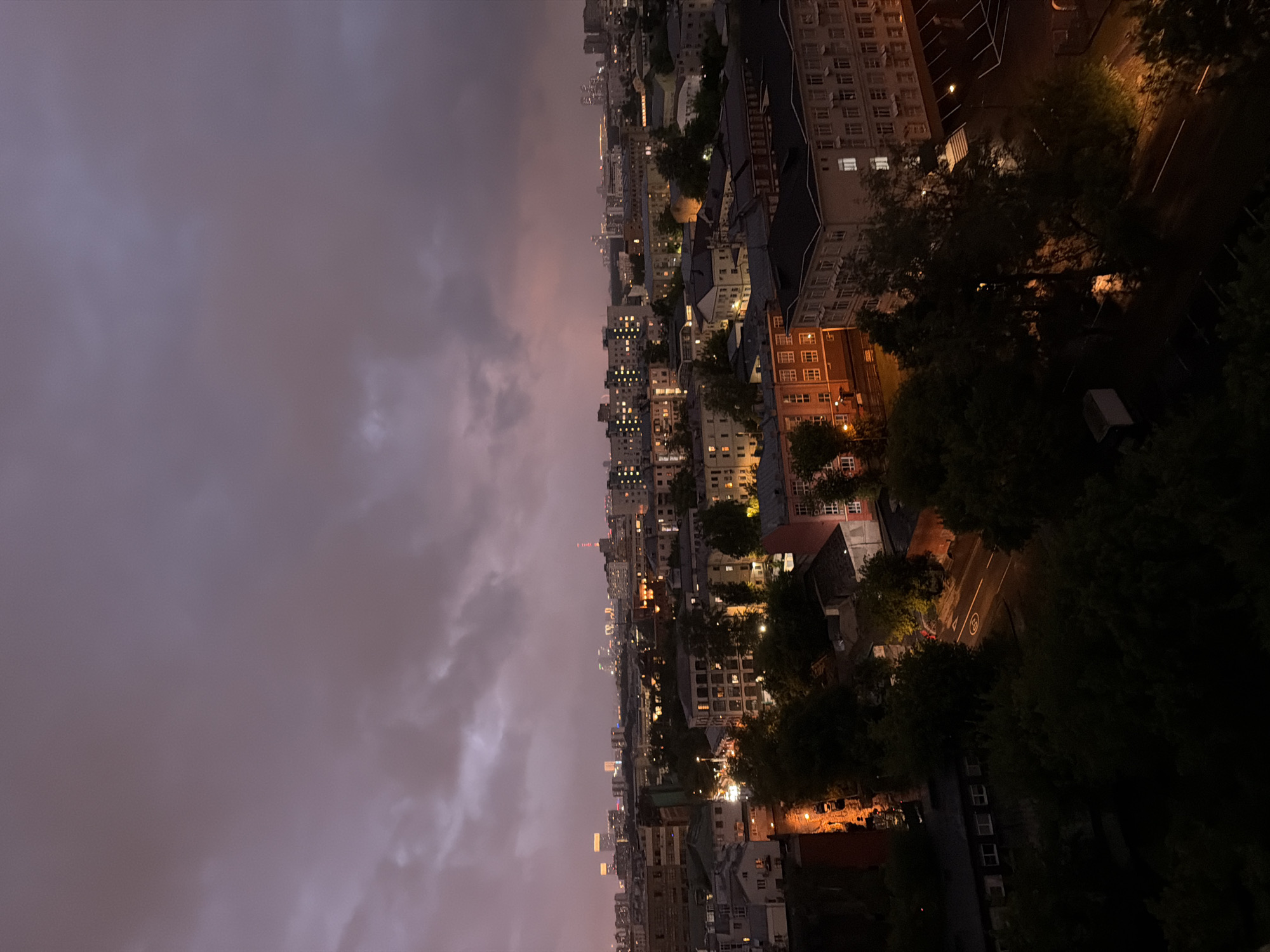 The image shows a cityscape at dusk with heavy, dark clouds overhead, suggesting an impending storm or recent rain. The sky is a muted purple and grey, with a hint of lighter color in the distance. Below the clouds, a dense urban environment is visible, with numerous buildings of varying heights and architectural styles. Many windows in the buildings are illuminated, indicating that it is evening. Trees are interspersed throughout the city, some appearing dark and full, while others are lighter and more sparse. A street is visible in the foreground, with cars parked along its side and streetlights casting pools of orange light. In the far distance, a skyline of taller buildings and city lights can be seen, suggesting a more modern part of the city. The overall impression is one of a slightly melancholic and atmospheric urban scene. The specific location is not identifiable without further information.