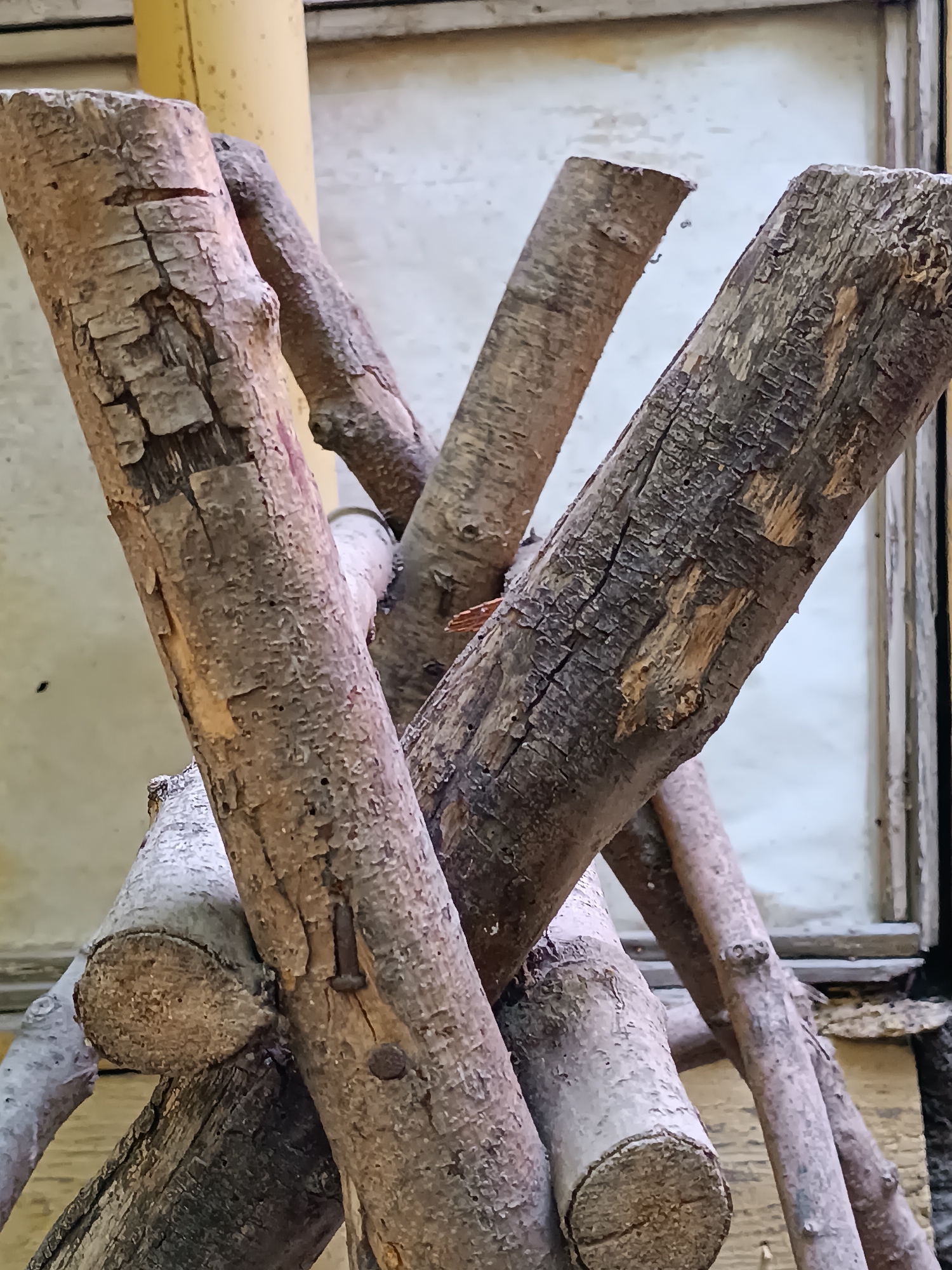 The image displays a close-up view of a rustic structure composed of several rough-hewn wooden branches. These branches, varying in thickness and texture, are artfully arranged in a cross-like configuration, with some appearing to be fastened together by visible nails. The wood exhibits a weathered appearance, with cracks and imperfections in its bark. In the background, a pale, textured surface is visible, possibly a wall or screen, and a hint of a yellow cylindrical object, likely a pole or pillar, is present. The overall impression is one of natural, handmade construction, likely an outdoor decorative element or support. The image does not contain any people or specific indications of location within Khashuri, Georgia.