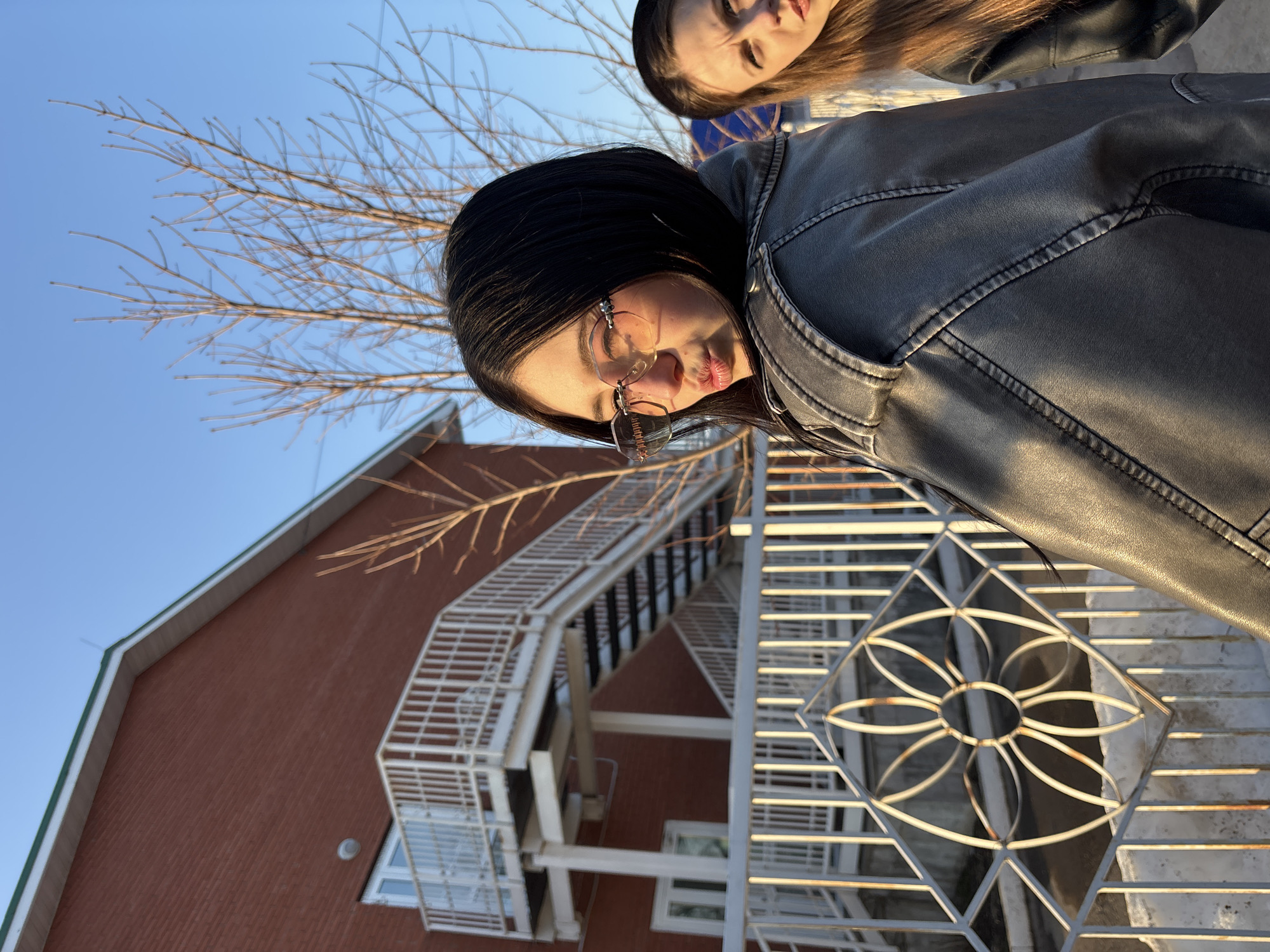The image depicts an outdoor scene in Фоки, Russia, featuring two young females in the foreground against a backdrop of a brick building and bare trees under a clear blue sky.
The primary subject, positioned to the left and closer to the viewer, is a young female with straight dark hair. She is turned over her left shoulder, looking towards the camera with her lips pursed. She wears rimless, lightly tinted glasses with decorative elements on the temples and a dark, possibly grey or faded black, leather-like jacket with prominent stitching. Her right ear has an earring.
Partially visible on the right, slightly behind the primary subject, is a second young female with long brown hair. She is looking upwards and to the right with a focused expression. Only her face, part of her hair, and her right shoulder, wearing a dark jacket, are visible.
In the background, a two-story building constructed of reddish-brown brick is visible. It features a prominent white metal external staircase with multiple landings and railings, attached to the building's facade. Several windows with white frames are visible on the building. The roofline has a light-colored fascia with a thin green trim. Bare deciduous tree branches are silhouetted against a bright, clear blue sky.
In the immediate foreground, a white metal fence or railing runs horizontally. It incorporates a decorative diamond-shaped panel with a floral or starburst pattern in its center, showing signs of light rust.
The scene is illuminated by strong, warm sunlight from the left, suggesting either early morning or late afternoon, which highlights textures and creates contrasts.
