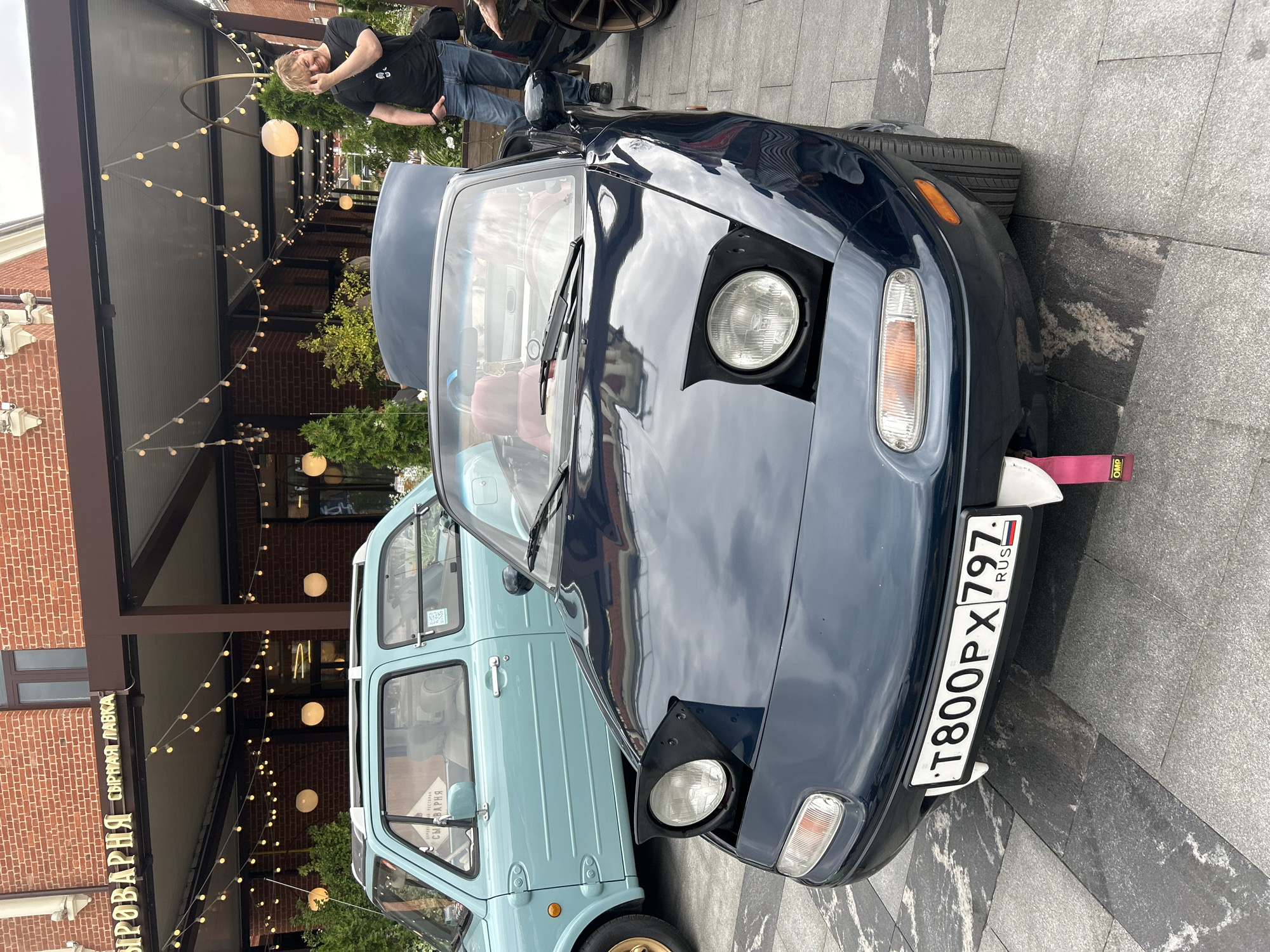 A dark blue convertible sports car, identified as a Mazda MX-5 Miata by its pop-up headlights and license plate "T800PX 797 RUS," is parked on a stone-tiled plaza. The car's hood is open, revealing a red interior. To the left, a light blue compact car, possibly a Nissan Figaro, is partially visible. Behind the cars, a building with a brick facade and a brown awning is adorned with strings of lights. A man in a black t-shirt and jeans stands to the right, gesturing towards his face. The scene is set in an outdoor area, likely a city street or public space, with trees and foliage present. The presence of the Russian license plate and the Cyrillic signage on the building indicates the location is Moscow, Russia.