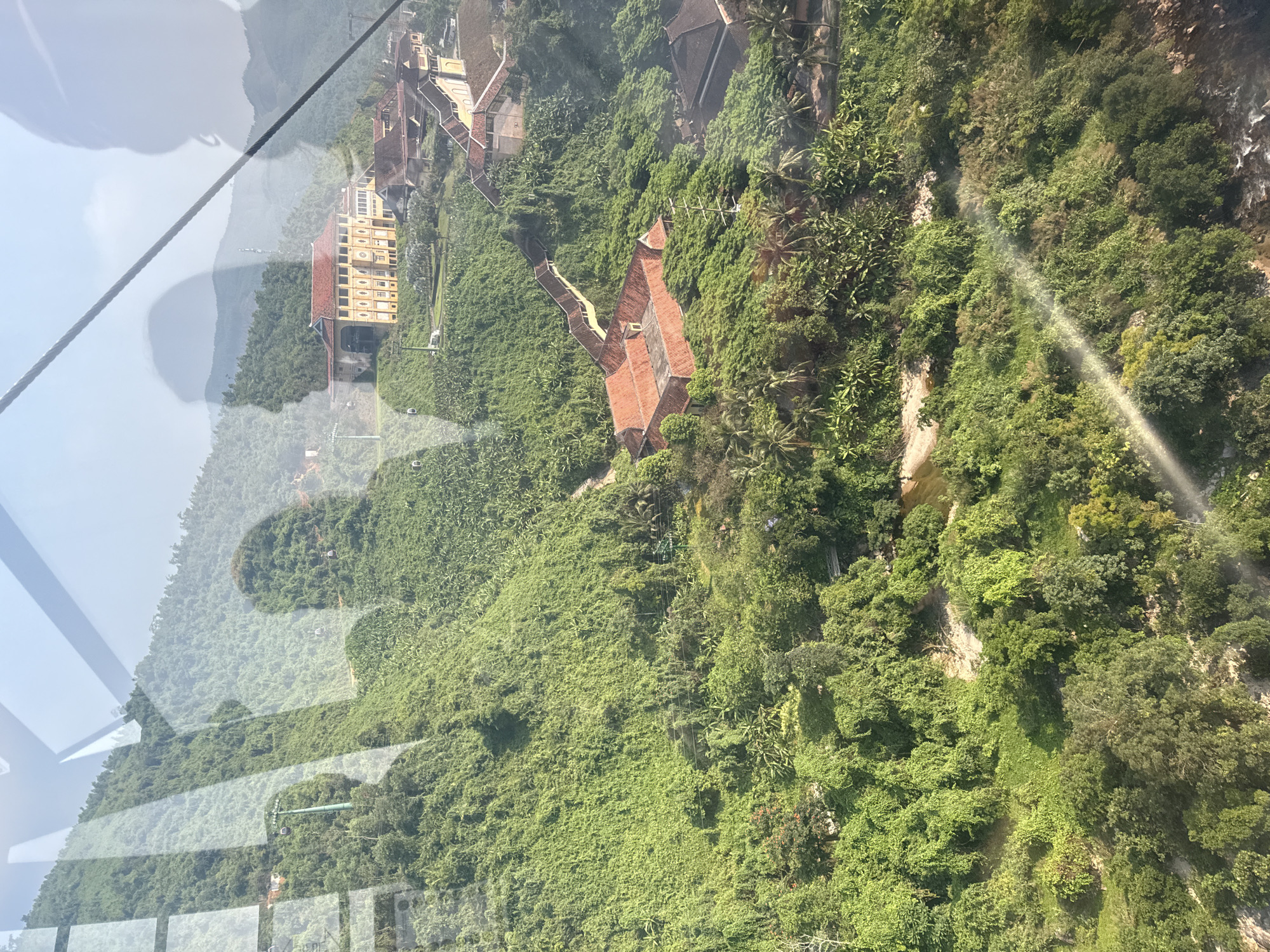 The image depicts a view from a cable car in Đà Nẵng, Vietnam, looking down upon a heavily vegetated mountainside. Buildings with red-tiled roofs are scattered among the trees. One building is a multi-story structure with a yellow facade. A faint reflection of the sky is visible in the foreground, and a cable car support cable crosses the upper left corner of the image. The dense vegetation appears lush and green, with hints of rock formations underneath. A small waterfall trickles down the hillside.