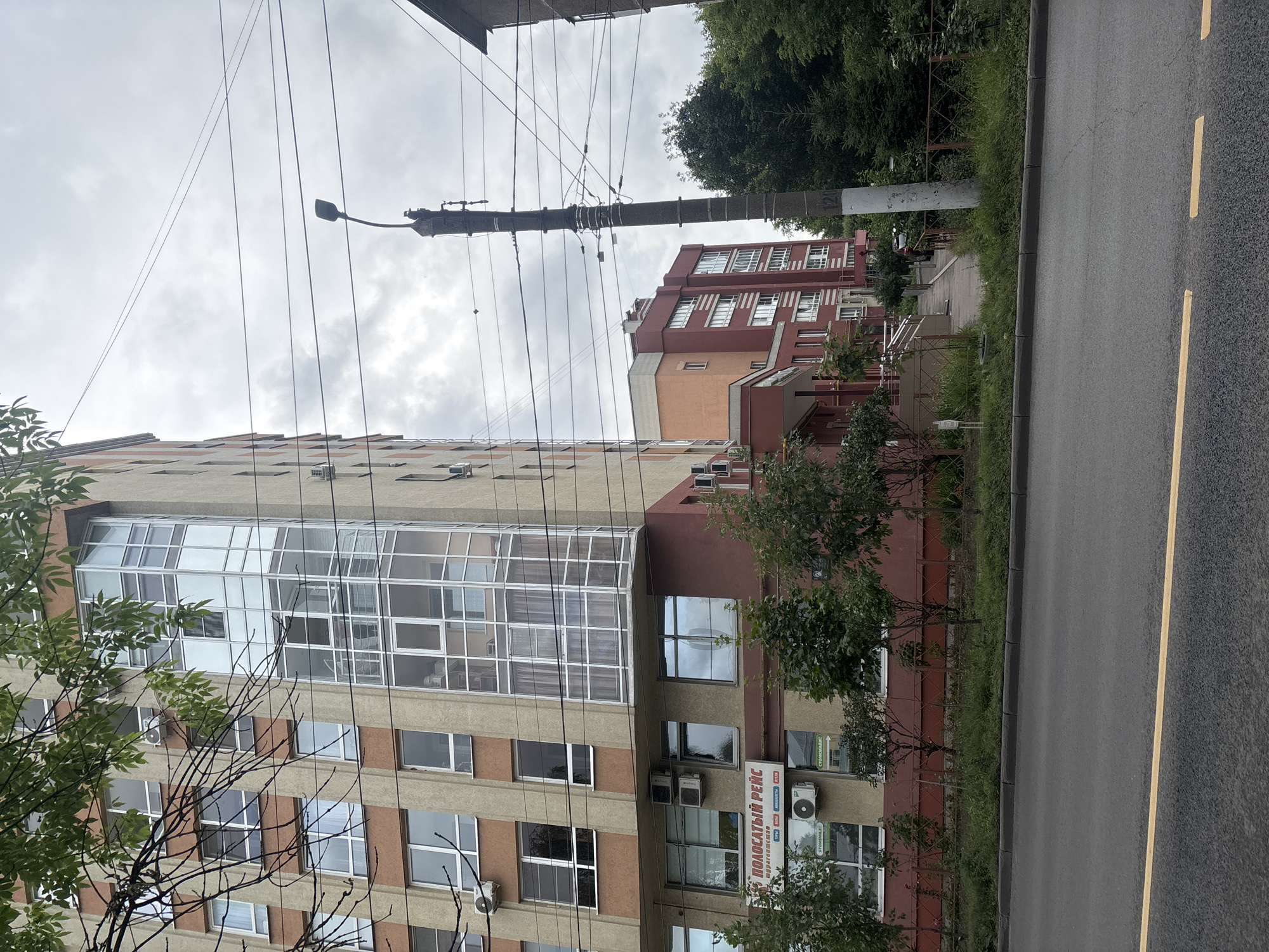 A tall, multi-story building with cream and brown colored facade is seen on the left. The building features numerous windows and a prominent glass-enclosed bay window section. A sign with Cyrillic text is visible on the lower part of the building. A sidewalk and road with a yellow line are in the foreground. A utility pole with electrical wires crosses the frame. In the background, another red and orange building is partially visible, along with trees and a cloudy sky. The scene appears to be an urban environment in Ivanovo, Russia.