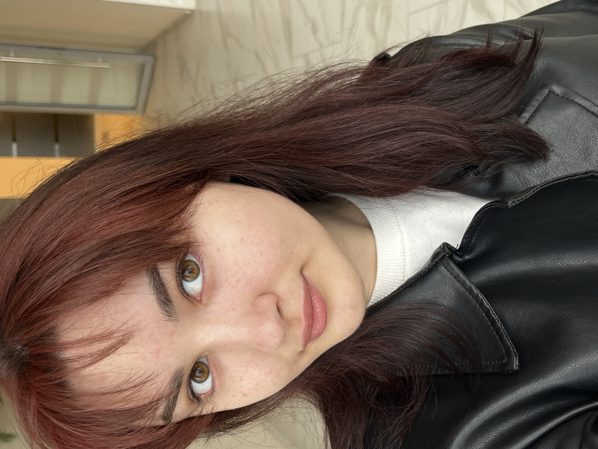 A close-up, angled portrait features a woman with reddish-brown hair, fair skin, and light brown eyes. She is wearing a white sweater under a black leather jacket. The background consists of a light-colored wall and what appears to be a shelf or cabinet. No information can be inferred about the location being in Neftekamsk, Russia from the provided image.