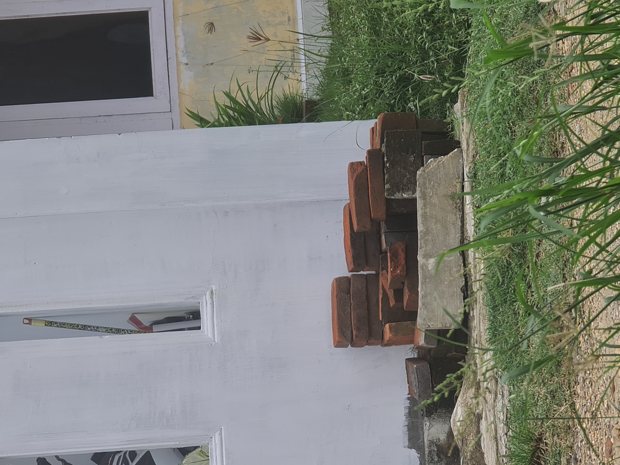 The image depicts an outdoor scene near a white building, possibly a house, in Lhokseumawe, Indonesia. A stack of red bricks and some grey concrete blocks are piled against the wall. In the background, lush green grass and a potted plant are visible. One of the windows of the building shows a broom with a patterned handle leaning inside. The overall impression is one of an exterior wall of a building with construction materials and vegetation surrounding it.