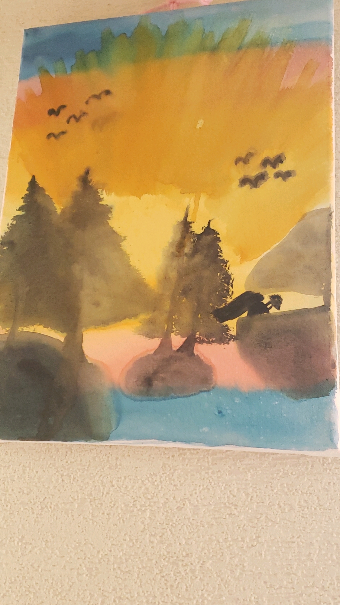 The image is a watercolor painting of a serene landscape at sunset. The sky transitions from a deep blue at the top to a vibrant orange and yellow, with hints of pink near the horizon. Silhouetted against the sky are several trees, some appearing as tall, dark spires, while others are smaller and clustered on what seems to be a small island. A body of water, painted in blues and greens, occupies the lower portion of the painting. In the distance, a few birds are depicted in flight, adding a sense of movement to the scene. The painting is hung on a textured beige wall. There is no discernible connection to the city of Prešov, Slovakia.