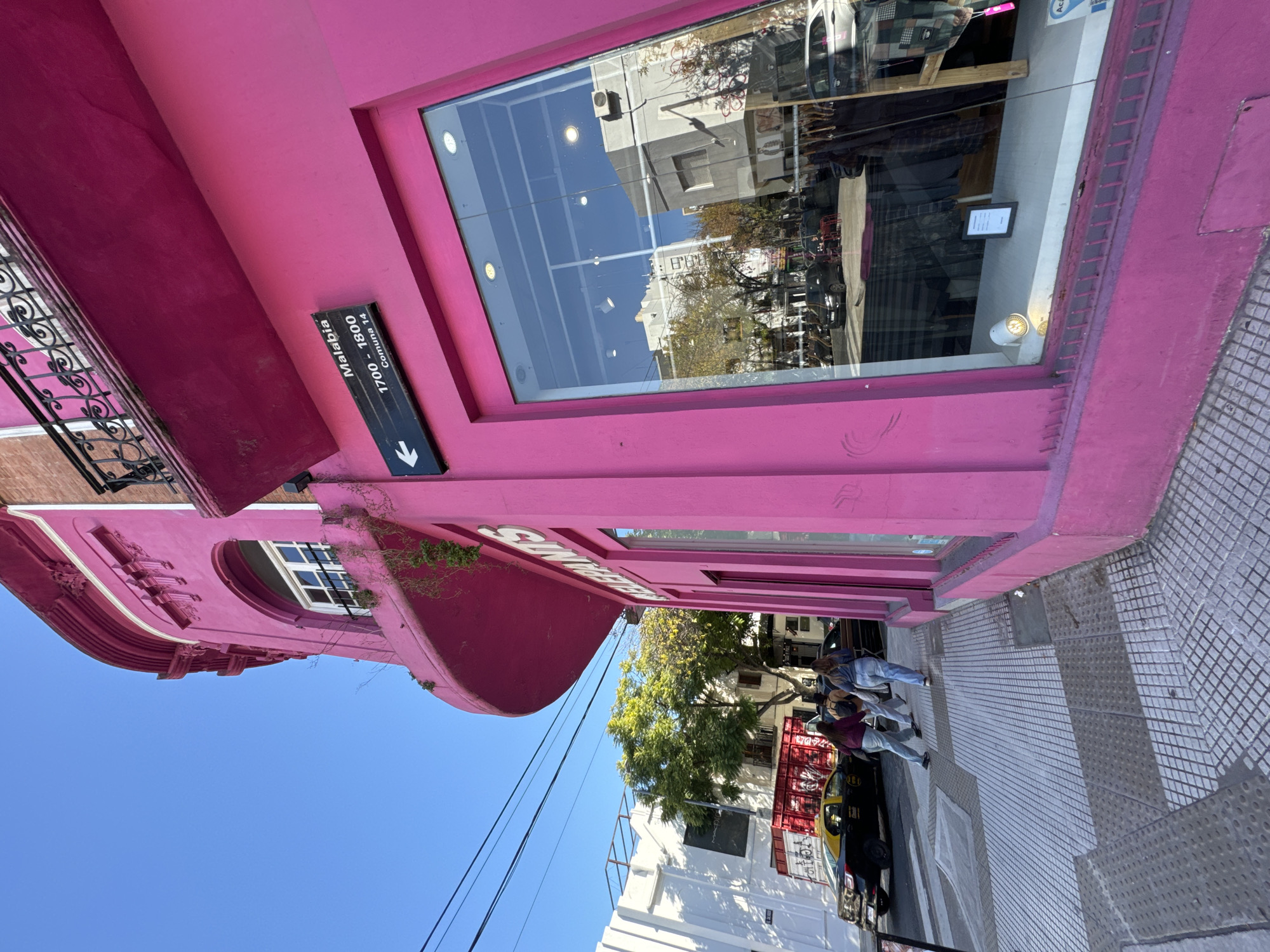 The image captures a pink building in Buenos Aires, Argentina. A street sign indicates "1700-1800 Malabia, Comuna 14". The building has a large storefront window reflecting the street. Two people are walking on the sidewalk in front of the building. A yellow and black taxi is parked nearby. The sky is clear and blue.