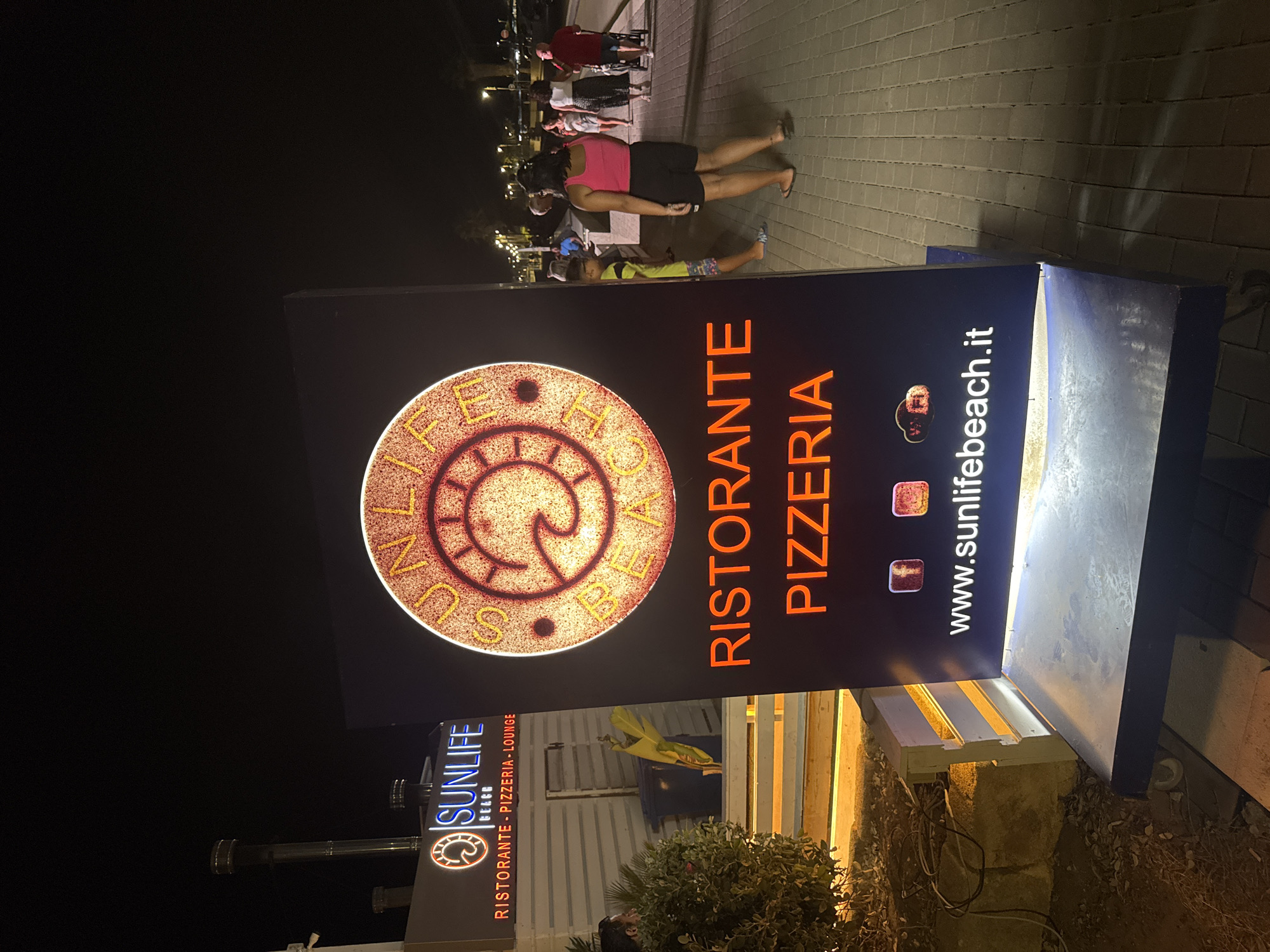 An outdoor freestanding illuminated sign for "Sunlife Beach" restaurant, pizzeria, and lounge is prominently displayed at night. The sign features a circular logo with a wave design and the words "Sunlife Beach" in orange neon. Below the logo, "RISTORANTE PIZZERIA" is written in large orange neon letters. At the bottom of the sign are icons for social media platforms (Facebook and Instagram) and the website "www.sunlifebeach.it". The sign is positioned on a wheeled base. In the background, to the left, a building with "SUNLIFE BEACH" signage is visible. To the right, a paved walkway with people walking is illuminated by streetlights. The location is Isola delle Femmine, Italy, as indicated by the presence of the sign which is associated with the Sunlife Beach establishment in that town. The ground is paved with light-colored bricks. Vegetation is present to the left of the sign.