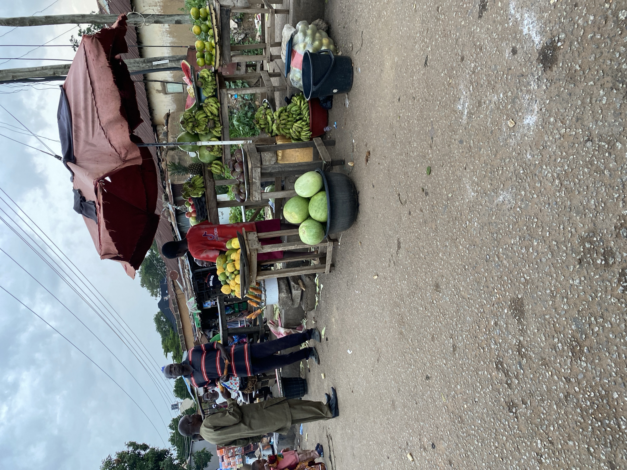 In the city of Bukuru, Nigeria, a bustling roadside fruit stand is captured under an overcast sky. Three men, one in a green kaftan and two in striped shirts, are engaged in transactions or conversations with vendors. The stand features a variety of produce, including bananas, watermelons, pineapples, oranges, and what appear to be corn on the cob. A large, worn, reddish-brown umbrella shields a portion of the stand from the elements. Wooden tables and crates display the colorful fruits, with black buckets and a plastic bag filled with oranges present on the ground. The scene is set on a rough, paved road, indicative of a typical urban market environment in the region.