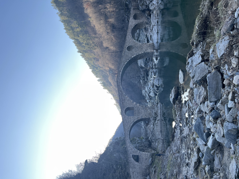 This image depicts the Dyavolski most (Devil's Bridge), a historic multi-arch stone bridge, spanning a river in a mountainous valley near Diadovtsi, Bulgaria. The bridge, constructed of grey masonry, features several arches over the water. The river's surface is calm, reflecting the bridge's arches and the clear, bright sky. The immediate foreground and riverbanks are covered with numerous grey and brown boulders and smaller stones, interspersed with patches of dry vegetation. One individual, dressed in dark attire, stands on the left riverbank near the easternmost arch of the bridge, facing away from the viewer towards the bridge or river. The valley is flanked by steep hillsides; the left slope is rocky with sparse, bare trees, while the right slope is densely forested with evergreen trees. A bright light source, likely the sun, is positioned in the upper left quadrant of the sky, creating a strong glare.