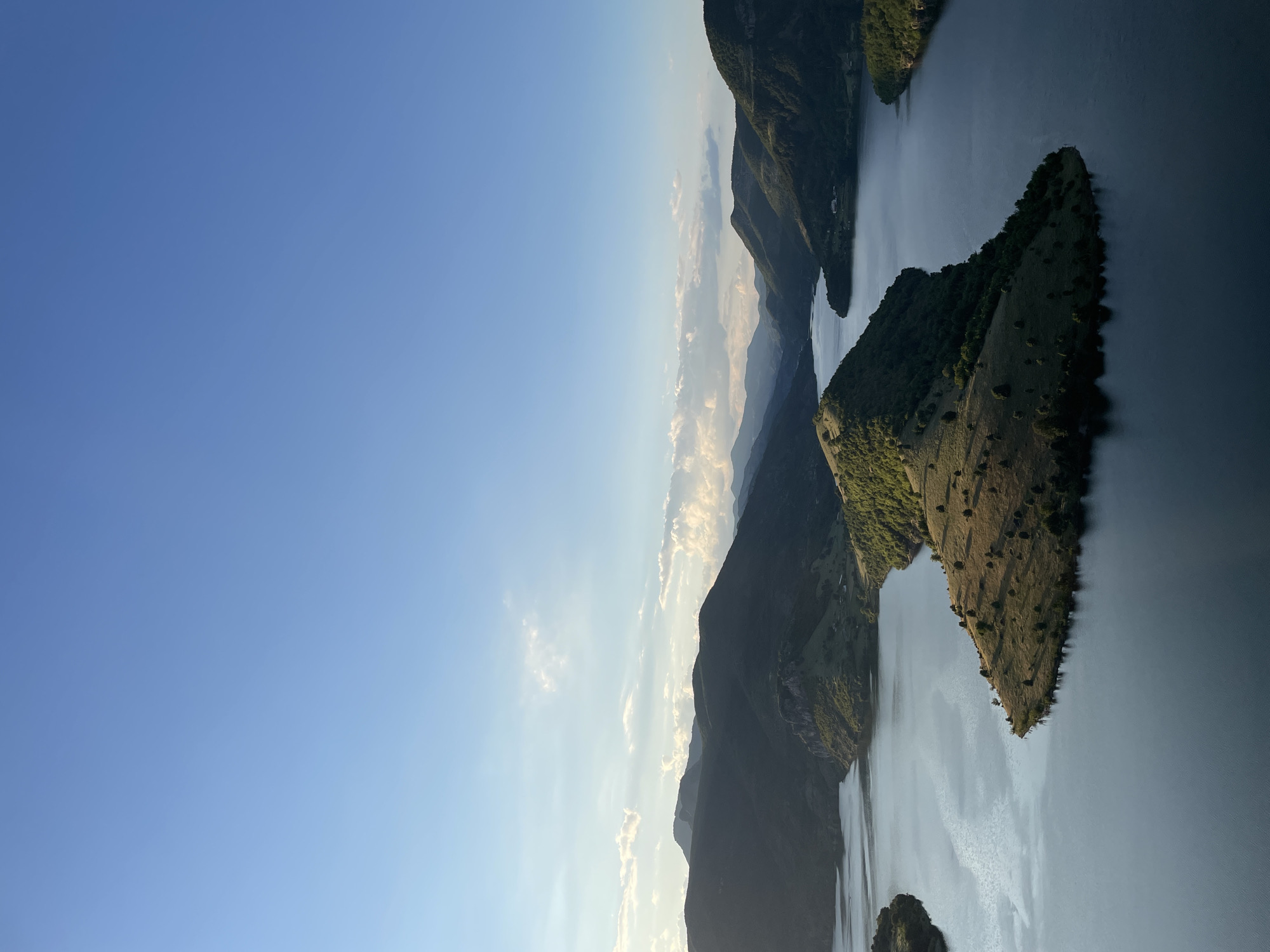 This is an aerial, wide-angle, landscape photograph of a serene lake nestled amongst rolling, verdant hills and distant mountains. The sky is a clear, pale blue with a scattering of wispy white clouds. The sun, low in the sky, casts a warm, golden light on the landscape, creating long shadows and highlighting the textures of the hillsides.
The foreground is dominated by the dark, still water of the lake, which reflects the sky and the surrounding terrain. A prominent, triangular peninsula juts out into the lake from the right side of the frame. This peninsula is covered in dry, golden grass dotted with scattered trees. Its slopes are gently contoured and bathed in sunlight.
To the left of the central peninsula, another, smaller section of the lake curves away, bordered by a hillside with less dense vegetation, allowing the water to be seen more clearly. Further into the background, the lake winds between increasingly dramatic mountain ranges, their peaks softened by distance and atmospheric haze. These mountains are covered in dark green foliage, appearing almost silhouetted against the brighter sky.
The overall impression is one of tranquil, natural beauty. The scene is devoid of any human presence or activity, emphasizing the untouched and peaceful nature of the location. The photograph appears to have been taken from a high vantage point, offering a sweeping vista of the lake and its surrounding topography.
This image depicts a section of the Arda River, specifically within the area of Ribartsi, Bulgaria. The prominent peninsula in the foreground is likely part of the man-made Arda 2 Reservoir, a popular destination for its scenic beauty and recreational opportunities. The surrounding landscape is characteristic of the Eastern Rhodope Mountains, known for their dramatic geological formations and lush vegetation.