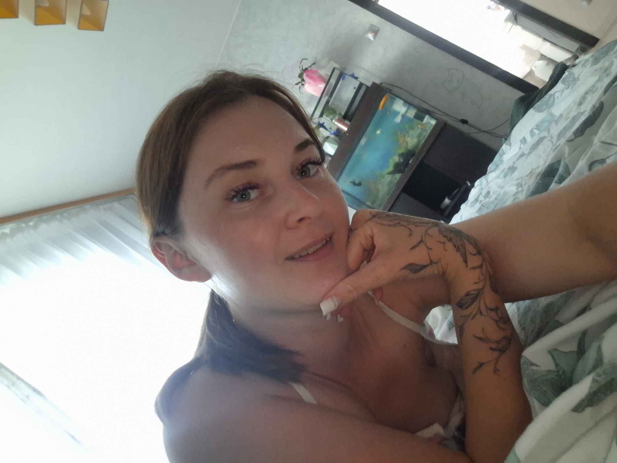 A woman is lying on a bed, looking at the camera. She has brown hair pulled back into a ponytail and is wearing a white bra. She is resting her chin on her hand, and her right arm is visible, adorned with a tattoo of birds and floral designs. To her left, a brightly lit window with sheer curtains is visible. In the background, an aquarium with fish and plants can be seen on a shelf, along with some decorative items. The overall lighting suggests it is daytime. The woman's expression is relaxed and she appears to be in a personal setting, possibly her bedroom. The location context is Марковский, Russia.