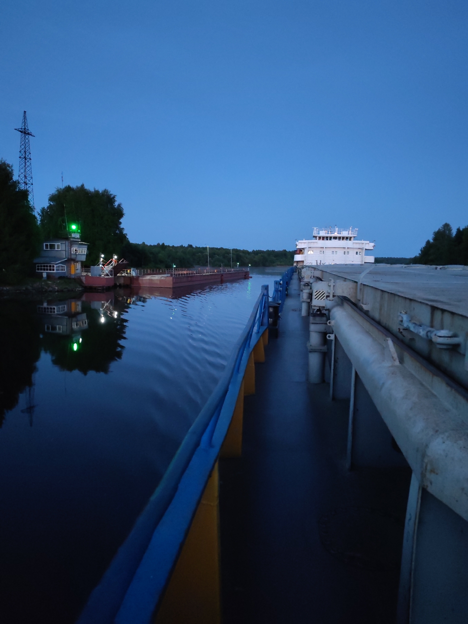 The image shows a view from the deck of a barge or ship on a dark blue body of water, likely a river or canal, at dusk. To the left, a red barge is moored near a small building with windows and a green light on top, which appears to be a control station or lock house. The building and a tall lattice tower are reflected in the calm water. In the distance, a white ship with multiple decks and windows is visible. The right side of the image is dominated by the deck of the vessel the photo is taken from, with a blue and yellow railing in the foreground. The sky is a deep, clear blue, indicating early evening or twilight. The scene is likely located in the vicinity of Annensky Most, Russia, a place known for its waterways and infrastructure. There are no people visible in the image.