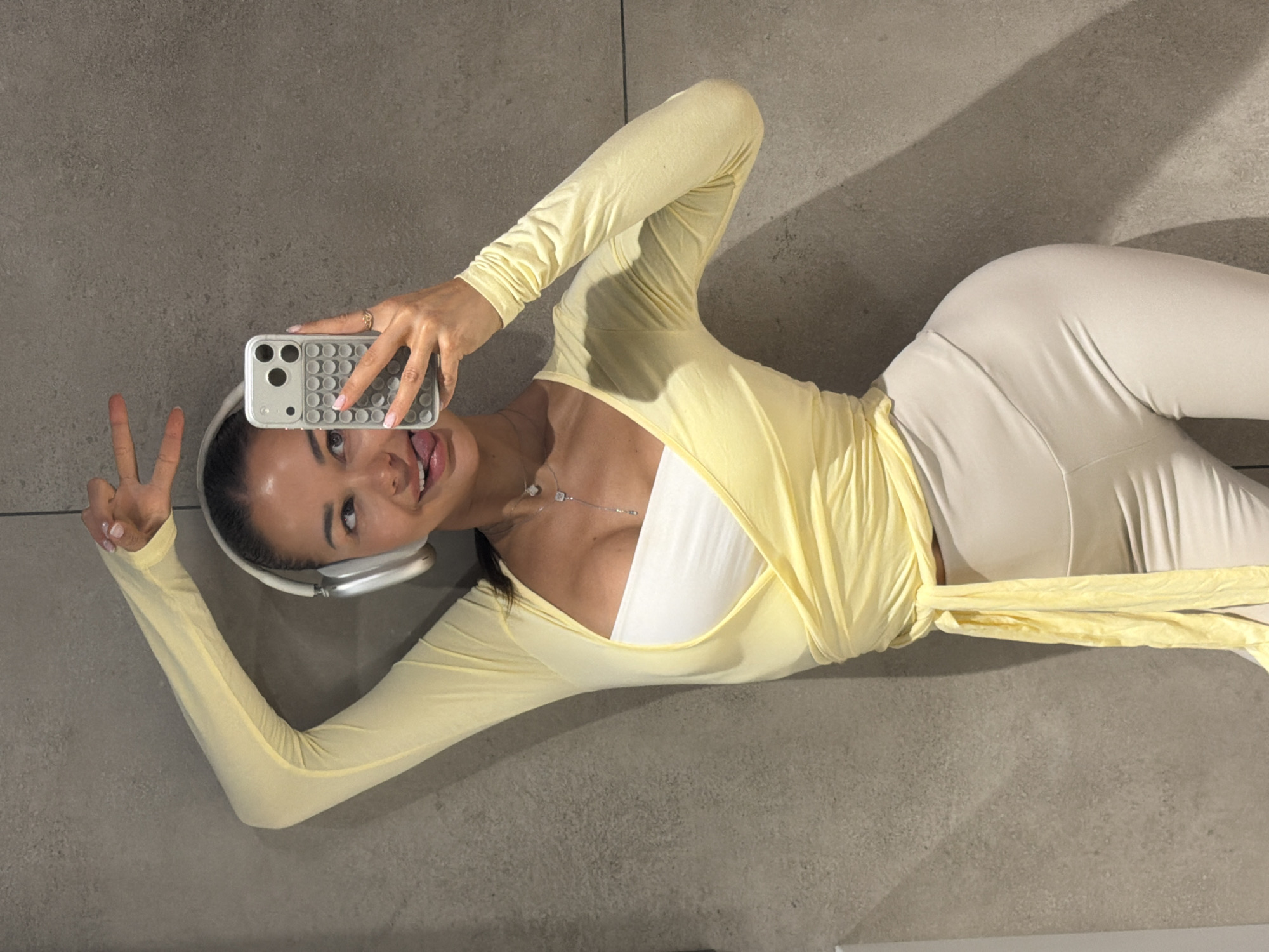 A young woman is lying on a light gray floor, taking a selfie with a smartphone, in what is described as Moscow, Russia. She is positioned diagonally across the frame, looking towards the camera with a playful expression, sticking her tongue out slightly and making a peace sign with her left hand.
She wears a form-fitting, light yellow long-sleeved top that wraps around her torso and ties at the waist, revealing a white bandeau or bra underneath. Her outfit is completed with light beige or off-white pants. She has a pair of silver and white over-ear headphones on her head and a delicate silver necklace with small pendants around her neck. Her right hand holds a light gray smartphone in a textured case, which features a grid of circular indentations, possibly resembling a 'pop-it' toy. Her fingernails are painted a light pink, and a ring is visible on her right ring finger.
The setting appears to be indoors, with a plain, light gray floor and wall creating the background. Strong, directional lighting casts distinct shadows from her body onto the floor, suggesting an artificial light source rather than natural daylight. The overall mood conveyed by her pose and expression is casual, cheerful, and confident. There is no visible text in the image.