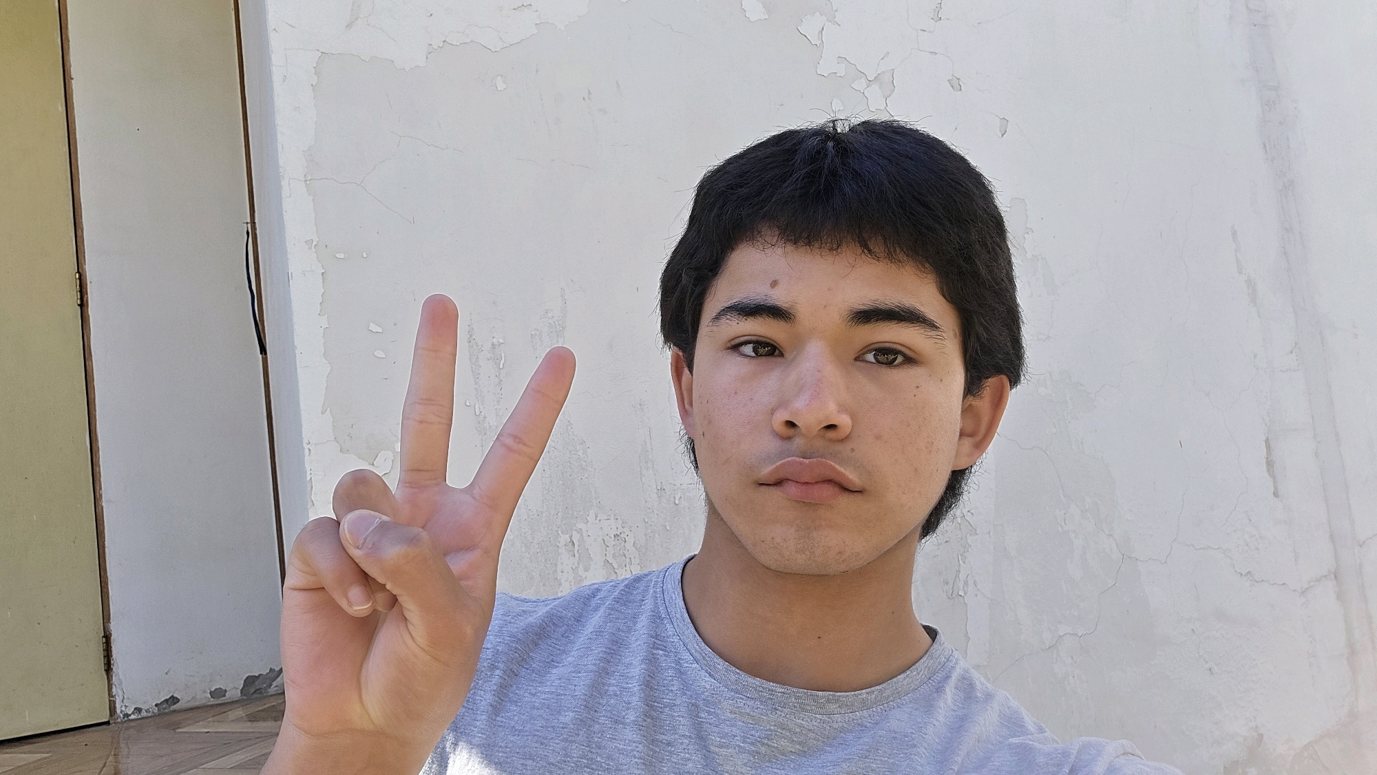 A young person with dark hair and brown eyes is pictured in a selfie. They are wearing a grey t-shirt and have their left hand raised, displaying a peace sign with their index and middle fingers extended. The background consists of a rough, off-white textured wall with some peeling paint, and to the left, a section of a beige door frame is visible, suggesting an outdoor or semi-outdoor setting. The lighting indicates it is daytime. The image does not contain enough information to determine its location in the city of Gultro, Chile.