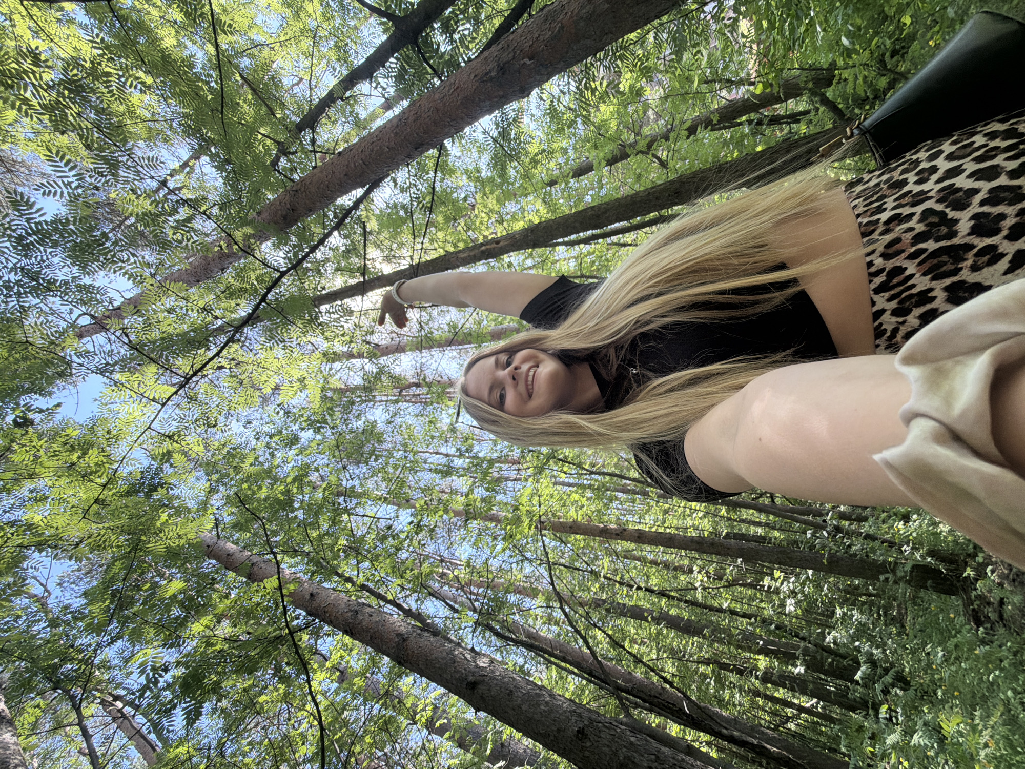 A woman is posing in a forest, looking up at the camera with her arm outstretched. She has long blonde hair and is wearing a black shirt and a leopard-print skirt. The background consists of tall trees and green foliage, with patches of blue sky visible through the canopy. The location is unspecified, so I cannot specify the location in the city of Izhevsk, Russia.