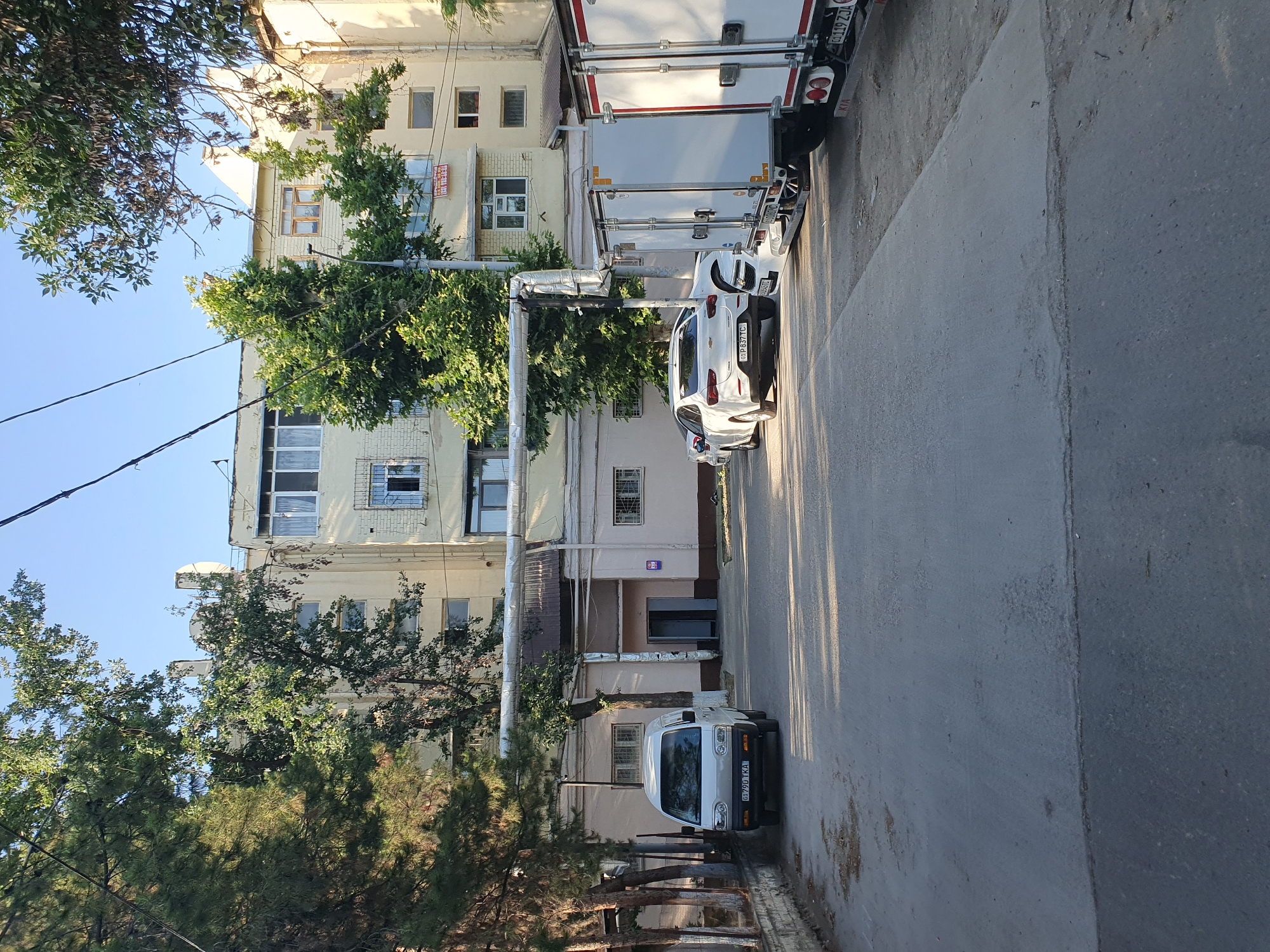 The image shows a street scene in Tashkent, Uzbekistan. In the foreground, a grey asphalt road runs towards a building. On the left side of the road, a white van with the license plate "01 790 TKA" is parked. Behind the van, there is a low building with windows and a dark-colored entrance. Further down the road, two white cars are parked, and to the right of these cars are three white box trucks.
The background features a multi-story building painted in a light yellow color. The building has numerous windows, some with bars, and exposed pipes running along its exterior. Green trees with lush foliage are visible on both sides of the street, casting shadows on the road. The sky above is clear and blue. A lamppost with a light fixture is also visible. There are no people or animals in the image. The scene appears to be a quiet street in a residential or commercial area.