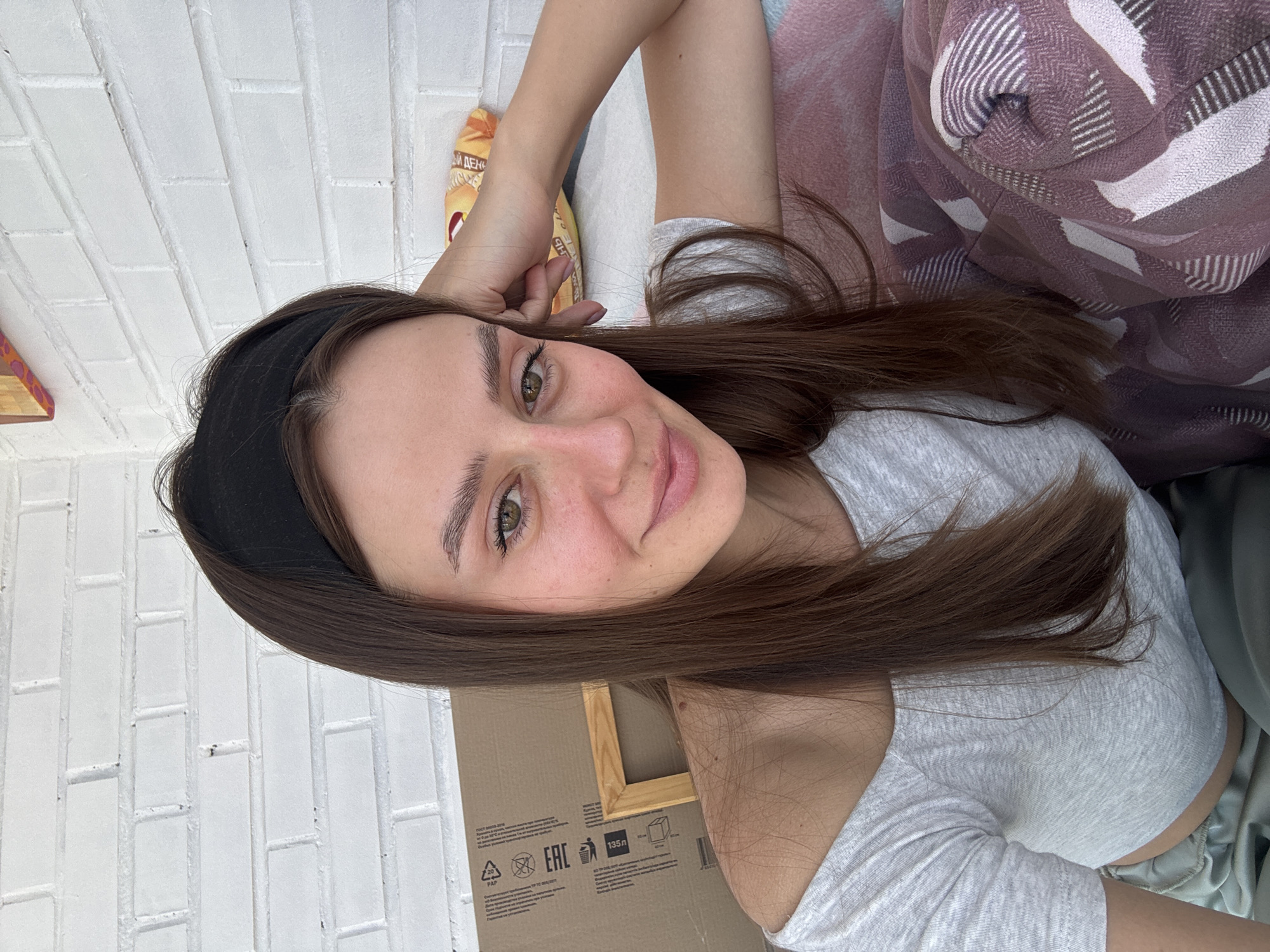 A young adult female is depicted in a close-up selfie, situated indoors or on a semi-enclosed balcony or porch in Krasnoyarsk, Russia. She has long, straight brown hair styled with a black headband, and green-hazel eyes. She is wearing a light grey off-the-shoulder crop top and partially visible light green trousers. Her right arm is bent with her elbow resting on a patterned purple and white blanket, while her head leans on her right hand. She is looking directly at the camera with a subtle smile. The background features a white painted brick wall. To her left, a large cardboard box with Cyrillic text and EAC (Eurasian Conformity) markings, indicating Russian origin, leans against the wall, with a wooden frame resting beside it. To her right, a yellow-orange snack bag with Cyrillic text is visible on the blanket.
