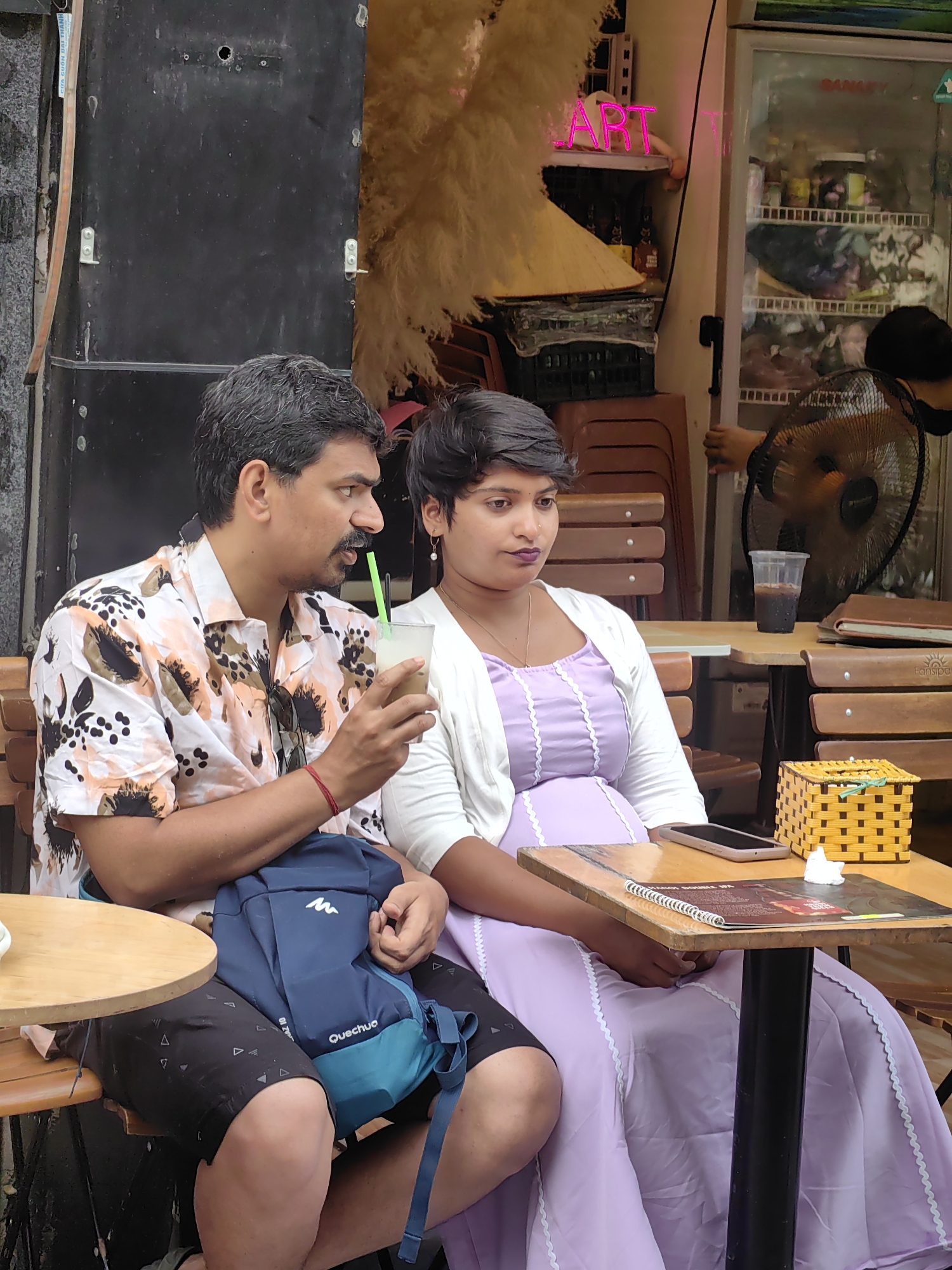 In Hà Nội, Vietnam, a man and a woman are seated at an outdoor cafe. The man, wearing a floral shirt and dark shorts, holds a drink with a green straw. A teal and blue backpack rests on his lap. The woman, wearing a lavender dress and white cardigan, sits next to him. A smartphone, a menu, and a yellow woven box are on the table. In the background are stacked chairs and a refrigerator with a pink "ART" sign. A person wearing a mask is visible behind a fan.