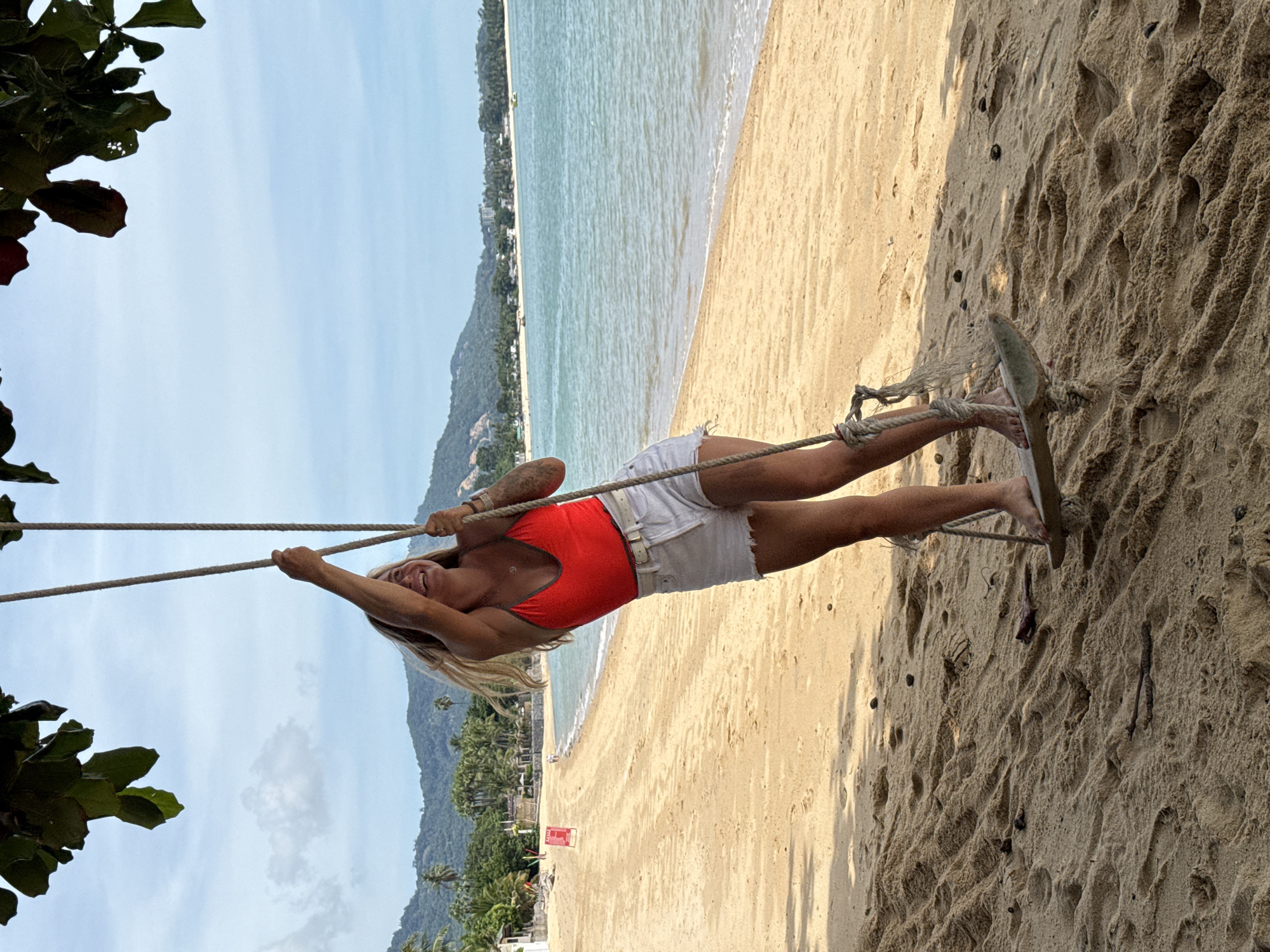 A young woman with blonde hair, wearing a red one-piece swimsuit and white denim shorts, is swinging on a rope swing on a sandy beach. The swing is suspended from a tree branch. The woman is smiling and looking upwards. The beach is golden sand and there is a calm, turquoise ocean in the background. Lush green hills with palm trees and some buildings are visible in the distance. The sky is light blue with some scattered clouds. The overall scene depicts a leisurely and happy moment at a tropical beach location, likely in or around Baan Bo Phut, Thailand.