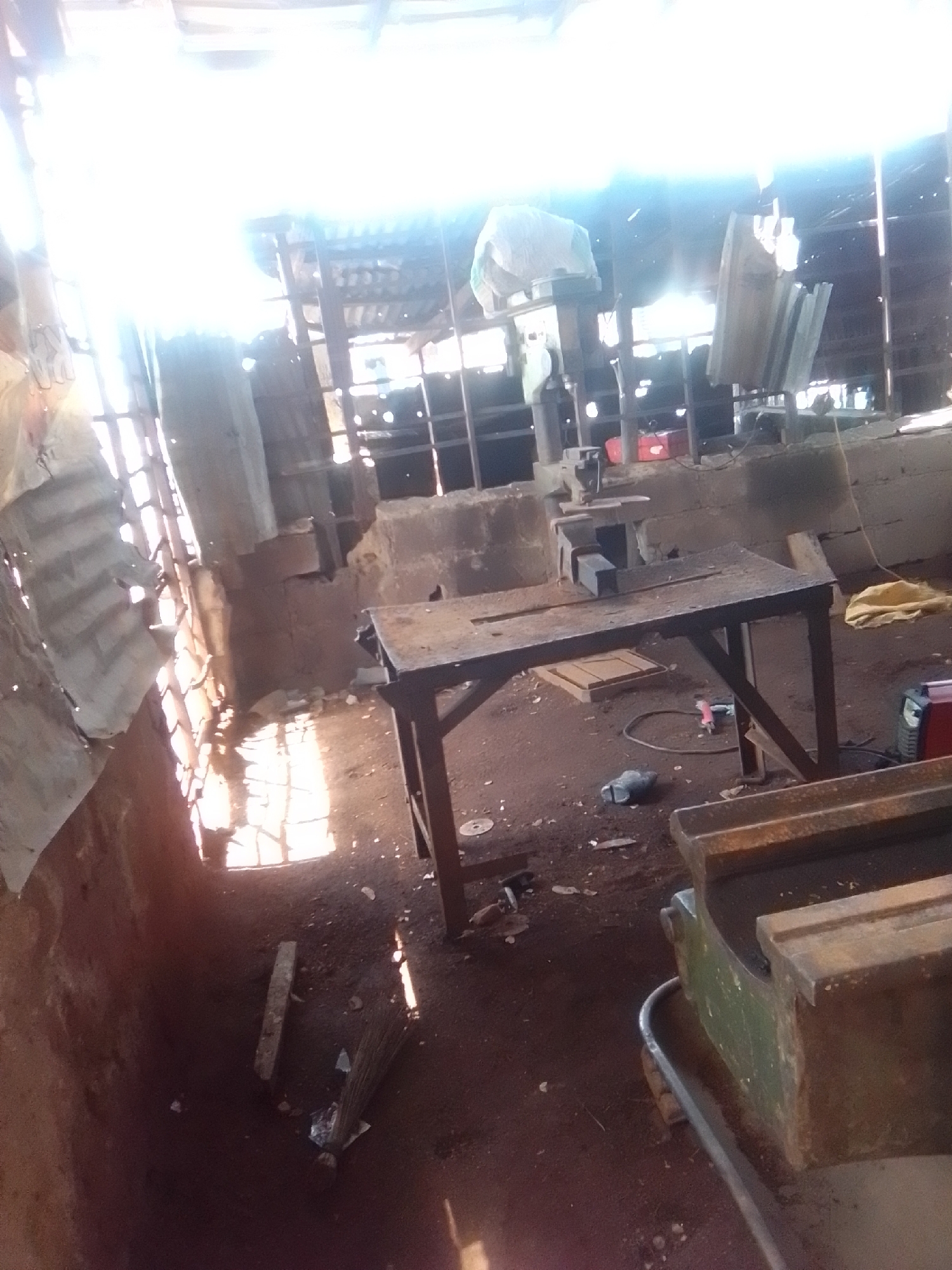 The image depicts the interior of a workshop, likely a metal fabrication or repair shop, situated in Bauchi, Nigeria. Sunlight streams in from the left through what appears to be a corrugated metal wall with open sections, illuminating the dusty, earthen floor. The workshop is cluttered with various tools and machinery.
In the foreground, on the right, a heavy, greenish-metal object, possibly part of a larger machine like a lathe or a press, rests on a metal frame. To the left of this, a workbench or table made of dark metal with a worn wooden or metal top is positioned. On this workbench, a vise with a metal clamp is visible, along with what looks like a piece of metal stock.
Scattered on the floor are tools and debris. A red electrical device, possibly a welding machine or power tool, is present towards the right side of the image, near a yellow piece of cloth or tarp. A coiled electrical cord or hose lies on the ground. Towards the back of the workshop, shelves and other industrial equipment are visible, partially obscured by shadows and the bright light. A broom stands upright near the left side of the image, suggesting ongoing cleaning or preparation for work. The overall impression is of a functional, albeit somewhat disorganized, working environment. There are no people clearly visible in the frame.