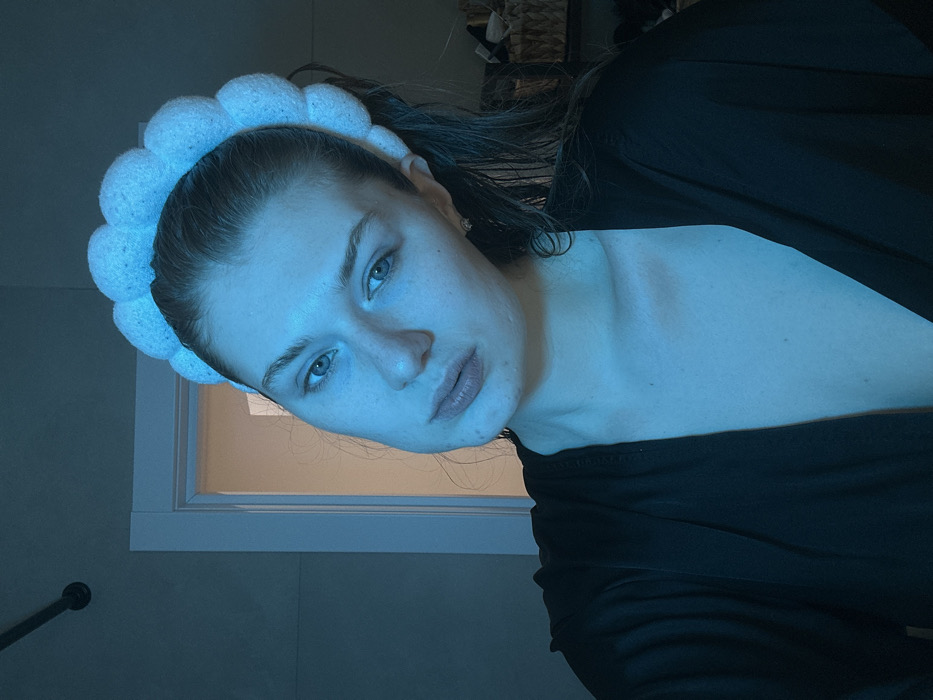 A young woman wearing a puffy white headband poses at a slight angle. She has fair skin, light eyes, and her hair is pulled back. She's wearing a dark top. The lighting is cool and blue. A window with light shining through is visible in the background. The scene appears to be indoors, potentially a bathroom, as a shower fixture is slightly visible in the lower left corner.