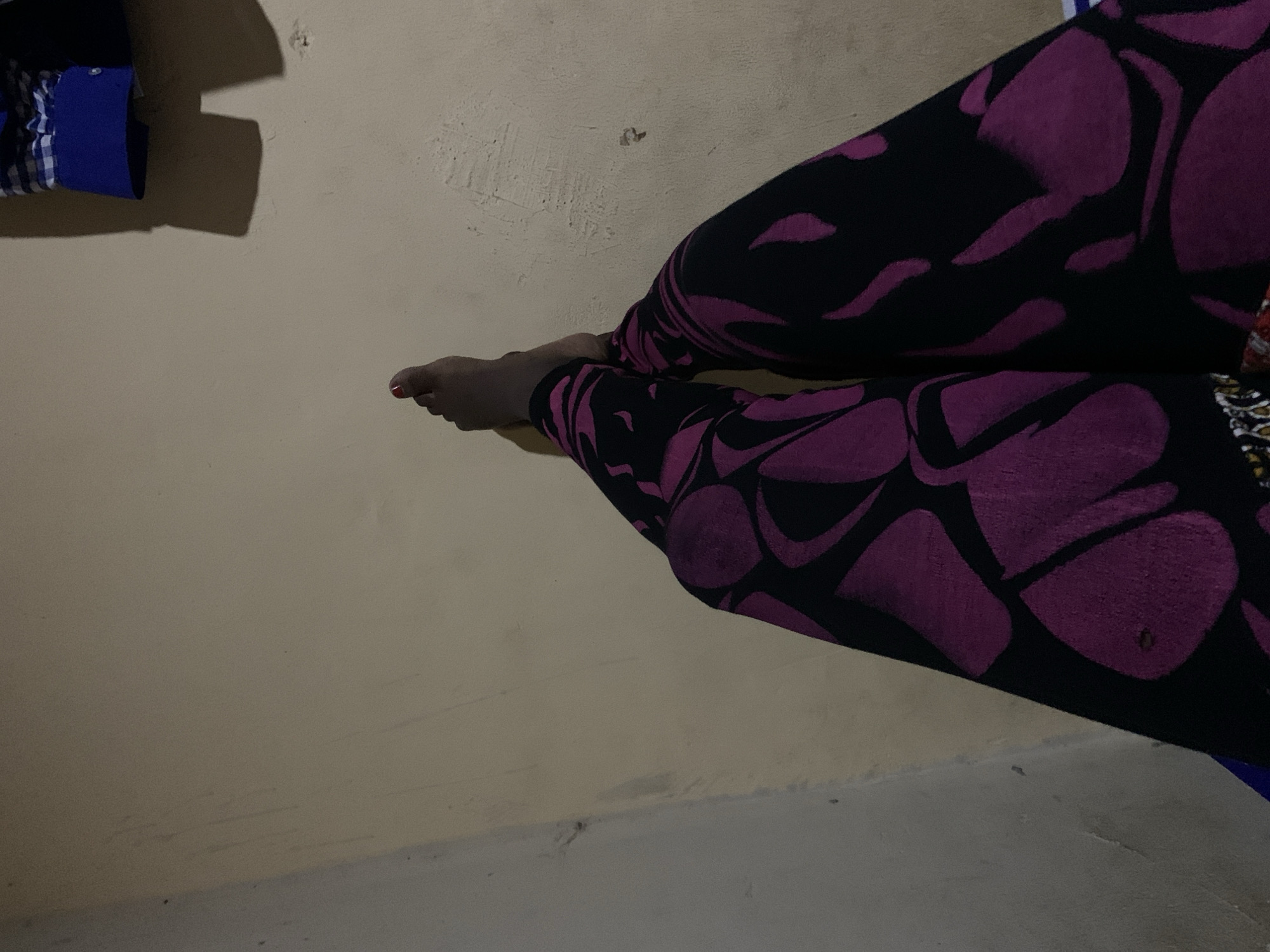 A person's legs are the primary focus of the image, positioned in the lower half and angled towards the right. The legs are covered by dark leggings adorned with a vibrant purple abstract pattern. One bare foot is visible, with red nail polish on the toenails. The background is a plain, textured wall in a neutral, light brown color. In the upper right corner, a folded blue and white checkered garment hangs, casting a shadow on the wall. The lighting suggests an indoor setting with artificial light, creating a soft illumination and some shadows. The scene is devoid of any contextual clues that would indicate a specific location within a city.