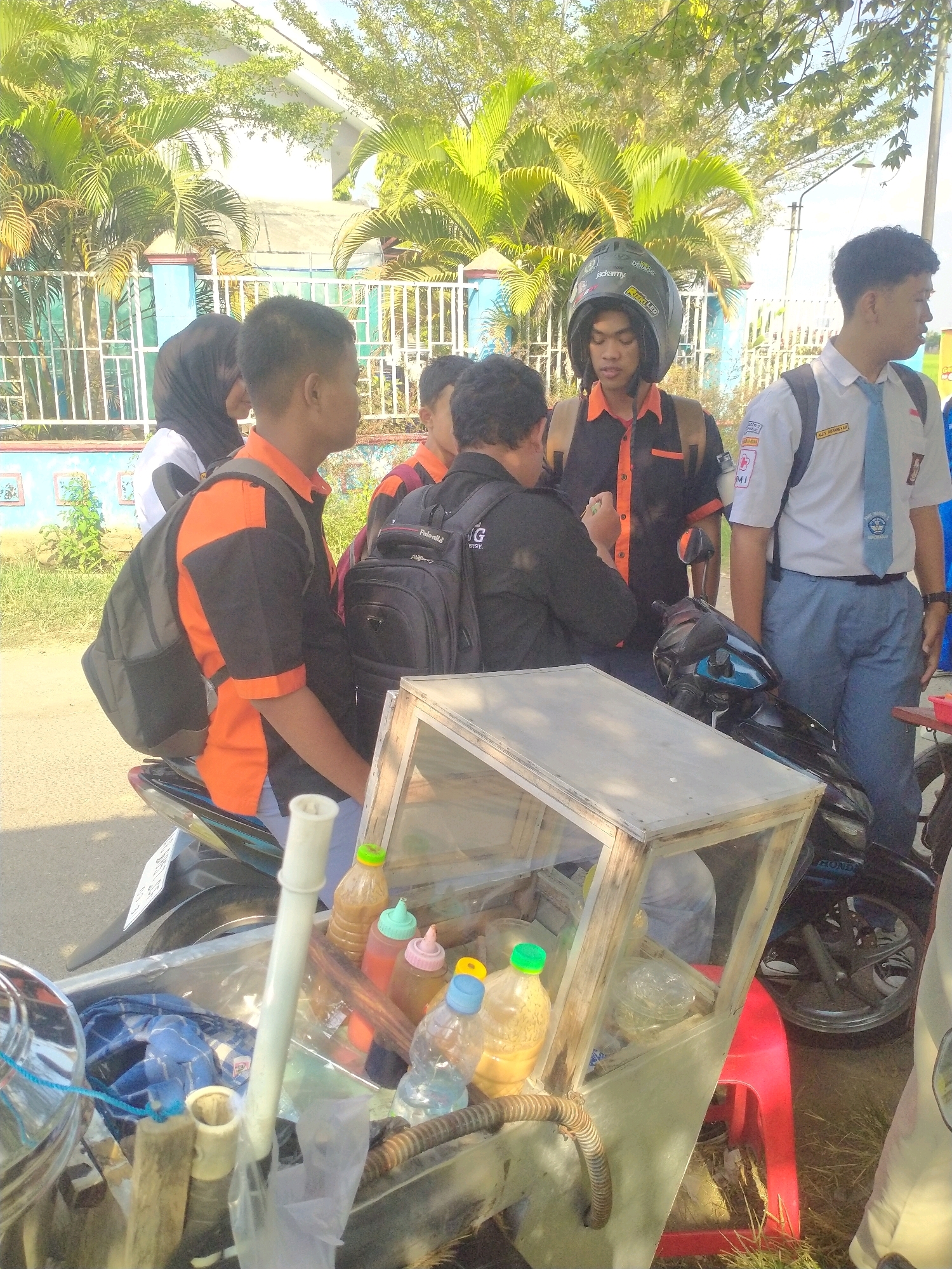 The image depicts a street scene in Begajah, Indonesia, on a sunny day. In the foreground, a makeshift food cart is visible, featuring bottles of condiments and a glass display case. A motorcycle is parked next to the cart, and several young people, likely students, are gathered around it. One individual, wearing a helmet and an orange and black shirt, is looking down. Another person in a black shirt and backpack is interacting with someone else. To the right, a person in a white school uniform with a blue tie is standing. Palm trees and foliage form the background, with a blue fence and a building visible behind them. The ground is paved.