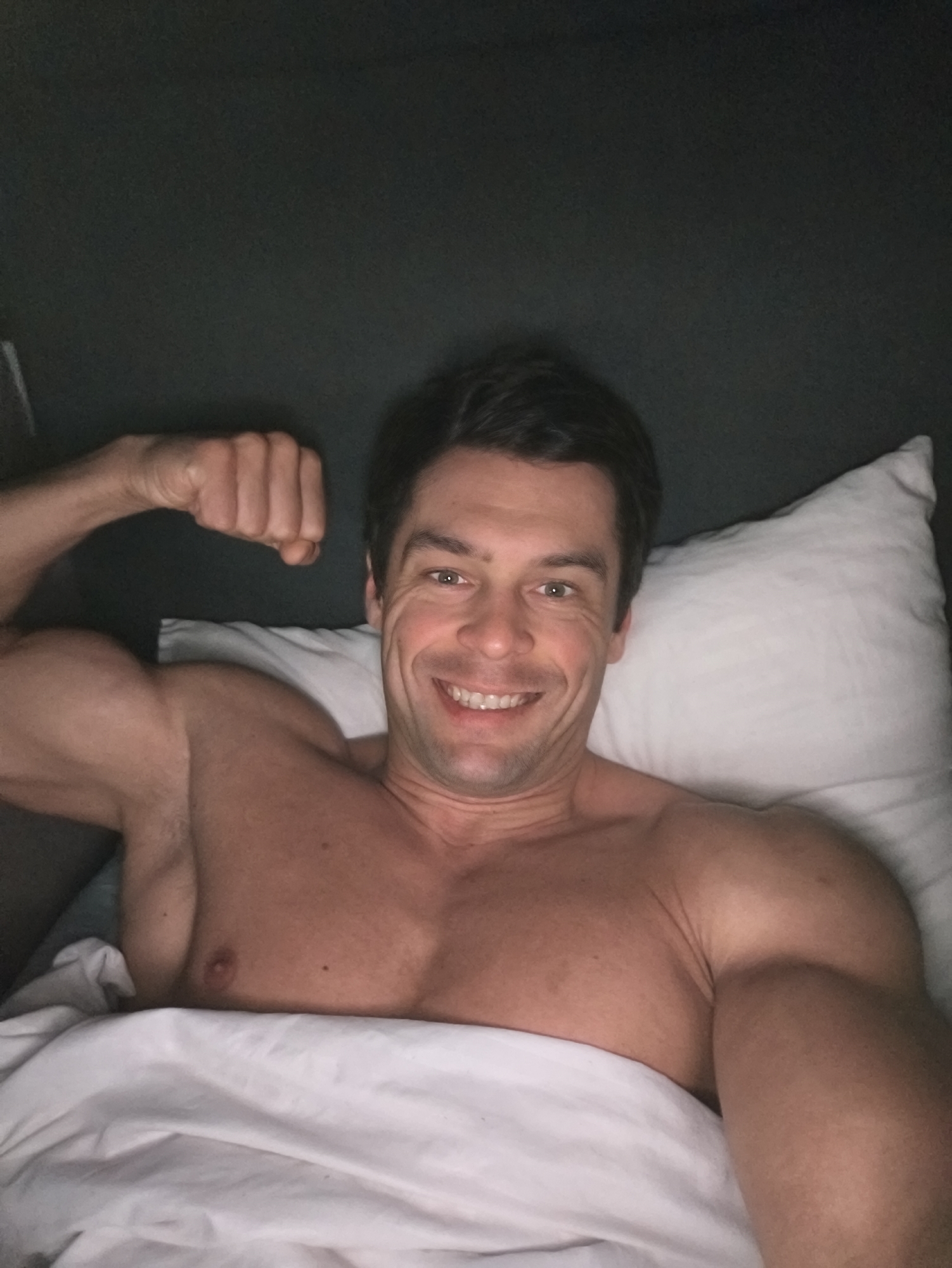 A shirtless man is lying in a bed with white sheets and a pillow. He is smiling and flexing his left bicep. The background appears to be a dark wall or headboard. The location is unknown and cannot be determined from the image.