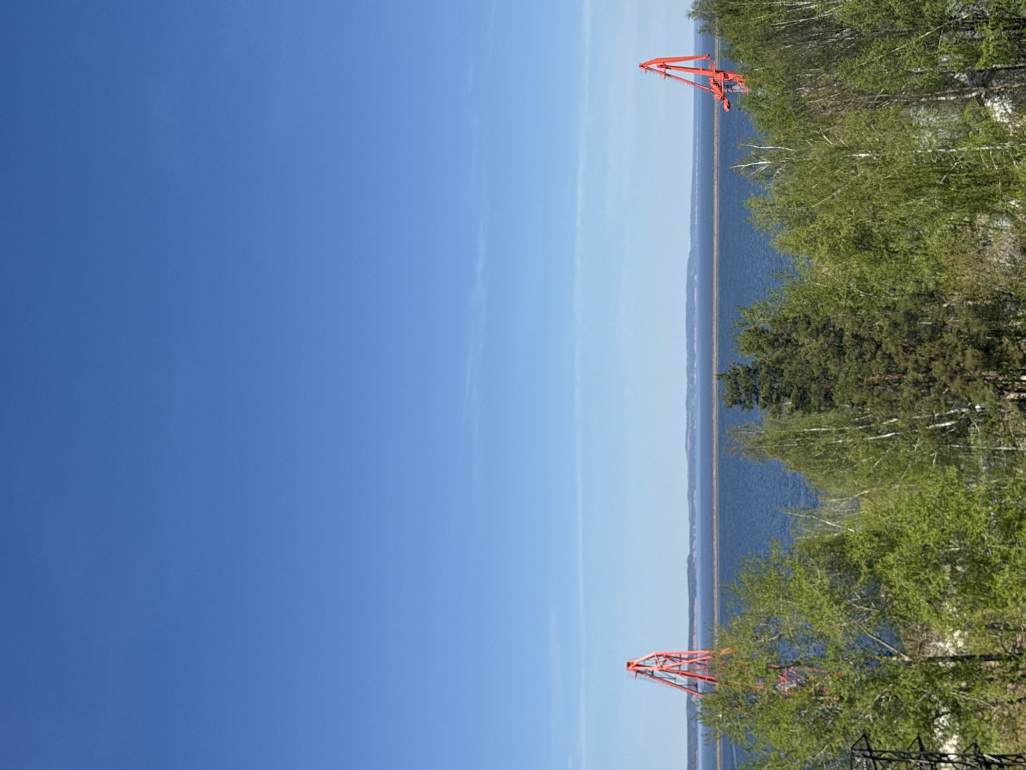 The image shows a view of the Kama Reservoir in Chaikovsky, Russia. The sky is clear blue with some scattered clouds. The reservoir is bordered by a forested area. Two orange, metal transmission towers are visible, one near the top of the frame and one near the bottom. The vegetation consists of a mix of deciduous and coniferous trees.