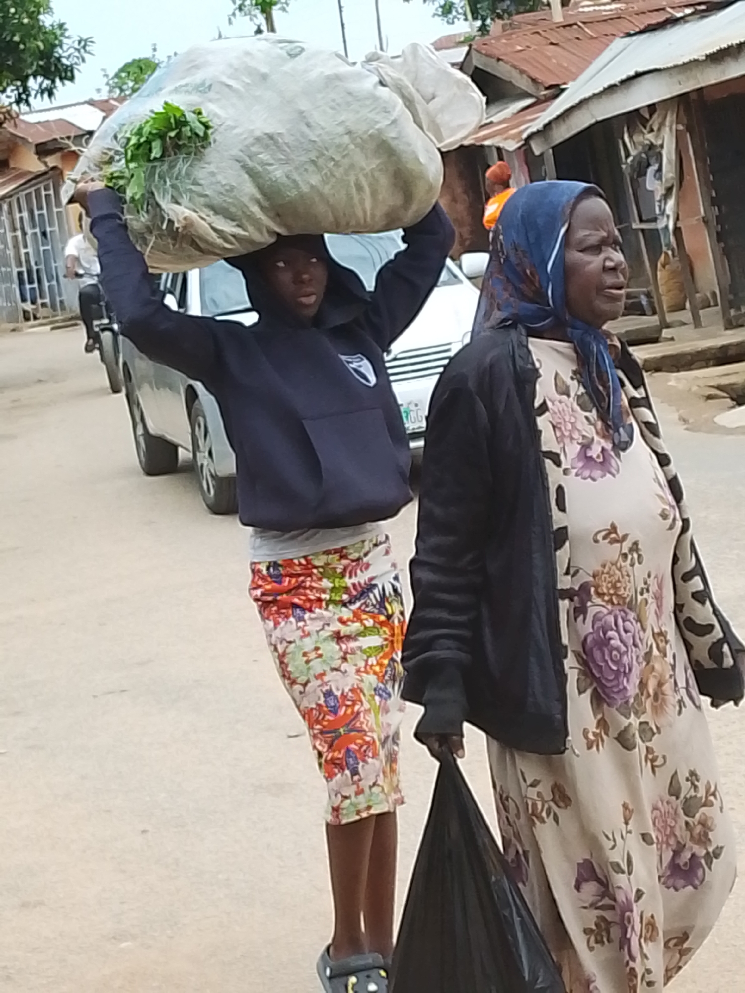 The image depicts a street scene in Jos, Nigeria. A young person wearing a navy blue hooded sweatshirt, a colorful floral skirt, and dark footwear carries a large white sack filled with leafy green vegetables on their head. They are walking alongside an older woman wearing a blue headscarf, a floral dress, and a dark jacket, who carries a black plastic bag. In the background, there are several parked cars, and behind them, buildings of varied construction, some appearing to have corrugated metal roofs. Other people are visible further in the background. The street is paved and appears to be in a residential area.