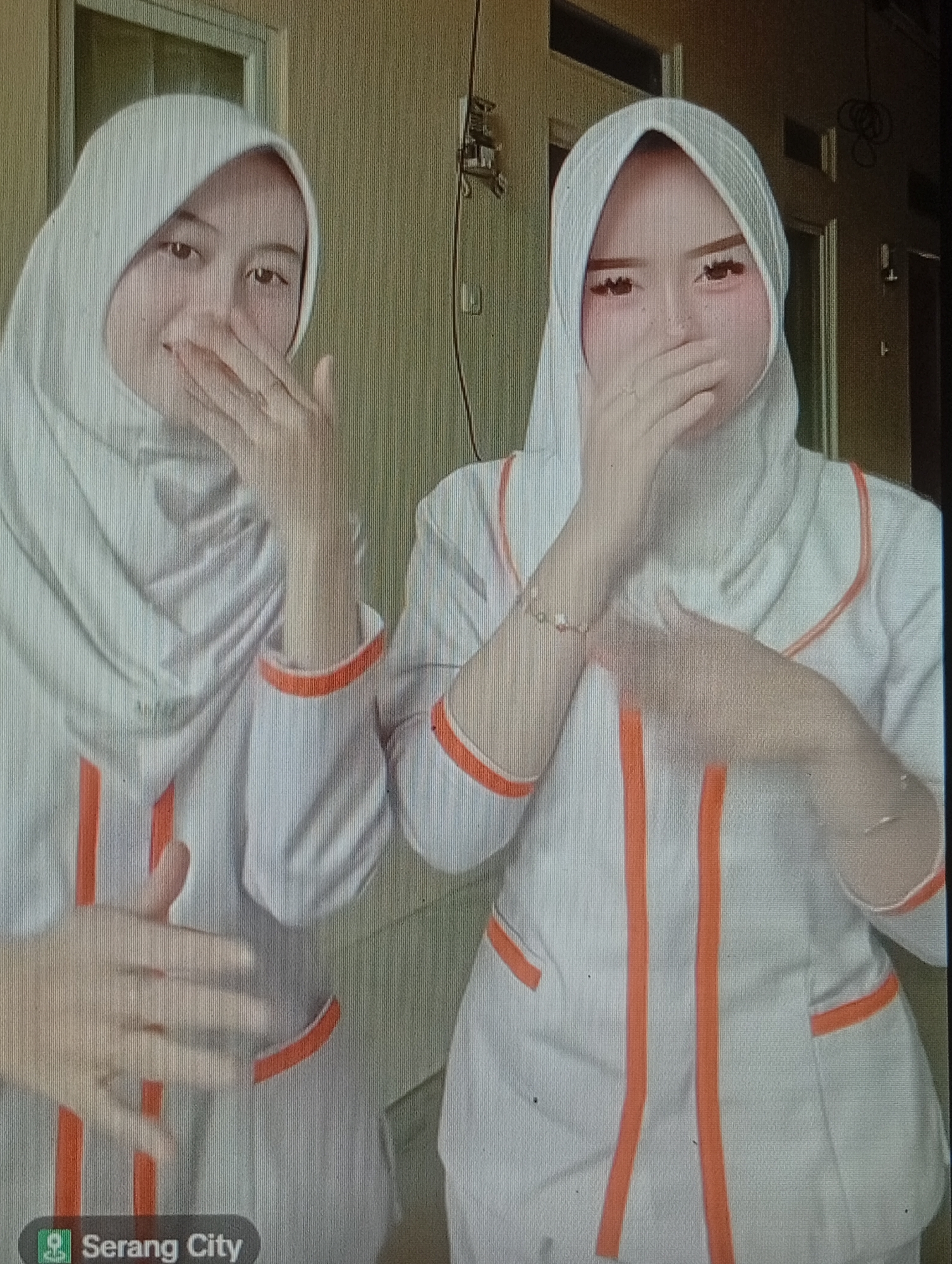 Two individuals wearing white hijabs and matching white uniforms with orange trim are posing for a photograph. The individual on the left has their hand covering their mouth, while the individual on the right has their hand partially covering their mouth and is wearing a bracelet on their left wrist. Both individuals appear to have makeup on. The background consists of a wall and doorways. A location tag in the bottom left corner indicates "Serang City," which is not in Lhokseumawe, Indonesia.