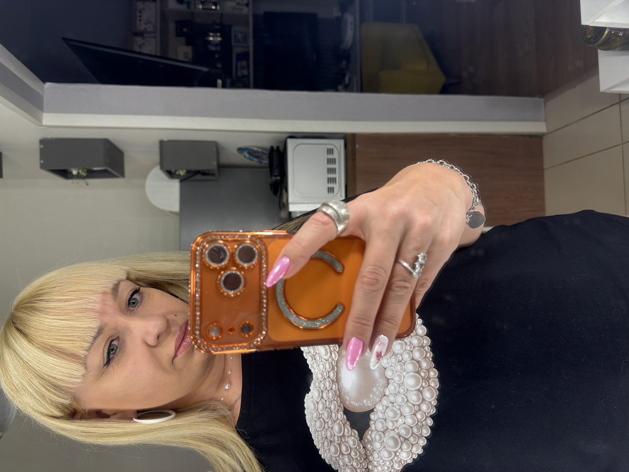 A woman with blonde hair and bangs is taking a selfie in what appears to be a home setting. She is holding an orange smartphone with a sparkly case in her right hand. Her nails are manicured with pink and white polish. She is wearing rings on her fingers and a silver charm bracelet on her wrist. She is wearing a black shirt with a large pearl or bead embellishment around the neckline. In the background, there is a shelf unit with various items on it, including a television and what appears to be a microwave. There are also square fixtures mounted on the ceiling. The reflection indicates she is in a room with neutral-colored walls and floors. The location in Chaikovsky, Russia is not evident in the image.