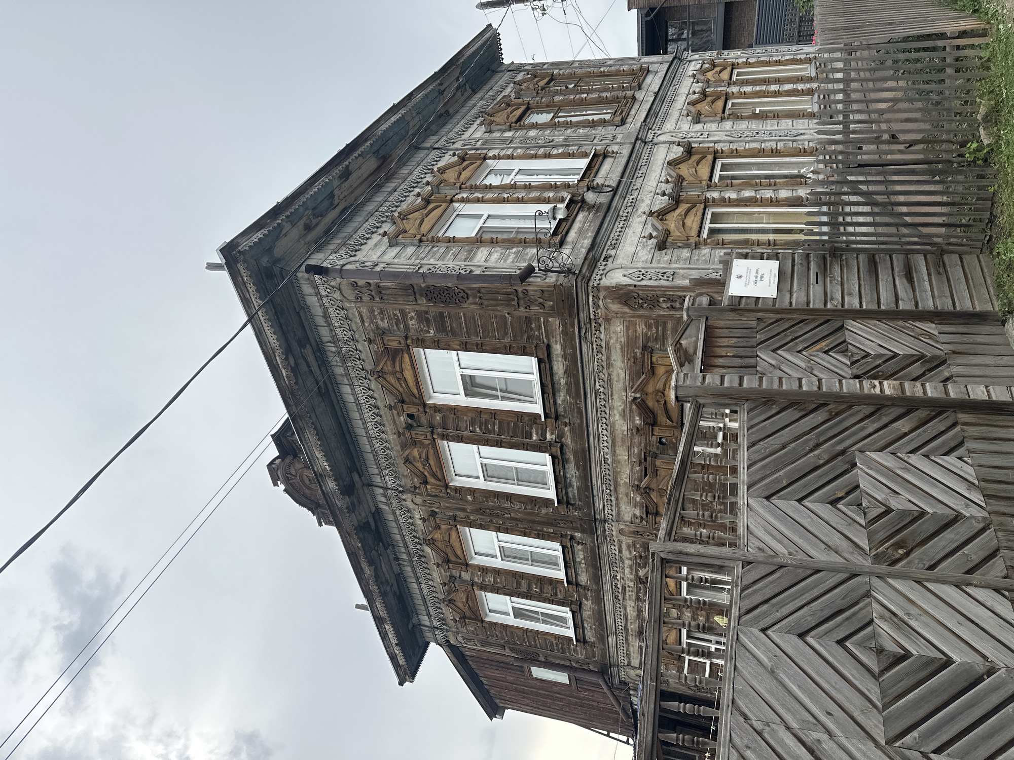 The image shows a two-story wooden house with ornate detailing, located in Biysk, Russia. The house features intricately carved window frames and decorative trim along the roofline. A wooden fence with a geometric pattern encloses the property. A sign attached to the fence indicates it is a historical building. The sky is overcast.