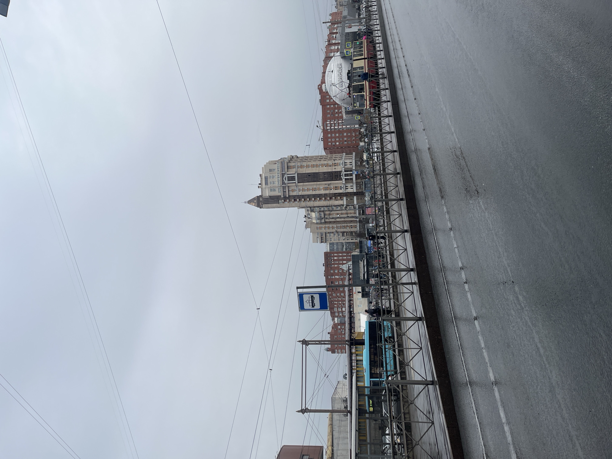 The image depicts a cityscape in Saint Petersburg, Russia, on a cloudy day. In the foreground, a tram stop is visible, marked with a blue sign. Behind the tram stop, a row of buildings dominates the background, including a tall building with a spire. Above the buildings, a large white dome-shaped structure stands out. Power lines extend across the sky. The street is wet, likely from rain. Several trams are visible. There are some people waiting at the tram stop.