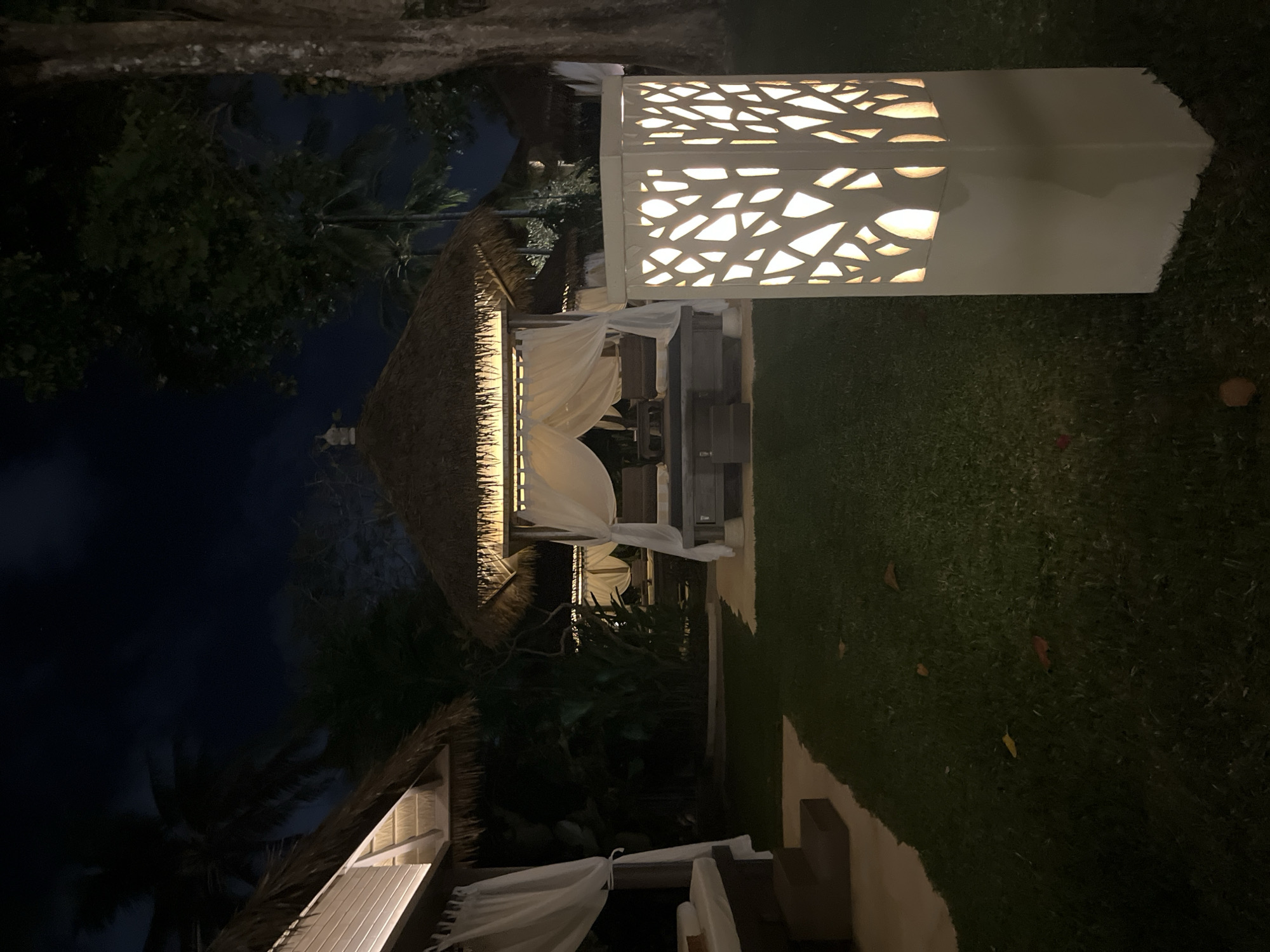 The image depicts an outdoor space, likely a resort or private garden, in Kuta Selatan, Indonesia, at night. The scene includes two thatched-roof gazebos adorned with white curtains. One is closer to the viewer, and the other is further back. A rectangular, illuminated decorative light fixture stands on the right side of the image, near the front gazebo. The area surrounding the structures appears to be well-maintained lawn. Dark sky and silhouettes of trees are visible in the background.
