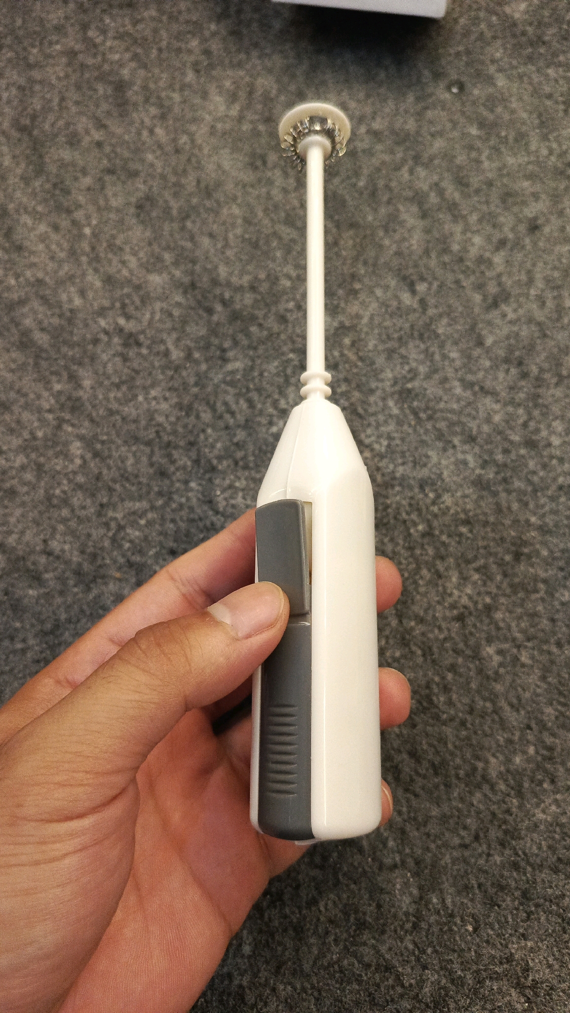 An overhead shot shows a person's hand holding a white and grey handheld milk frother. The frother has a long white stem with a circular, whisk-like attachment at the top. A grey rectangular button is visible on the side of the frother's white handle. The background is a grey, textured carpet. There is no indication of Babadan, Indonesia in the image.