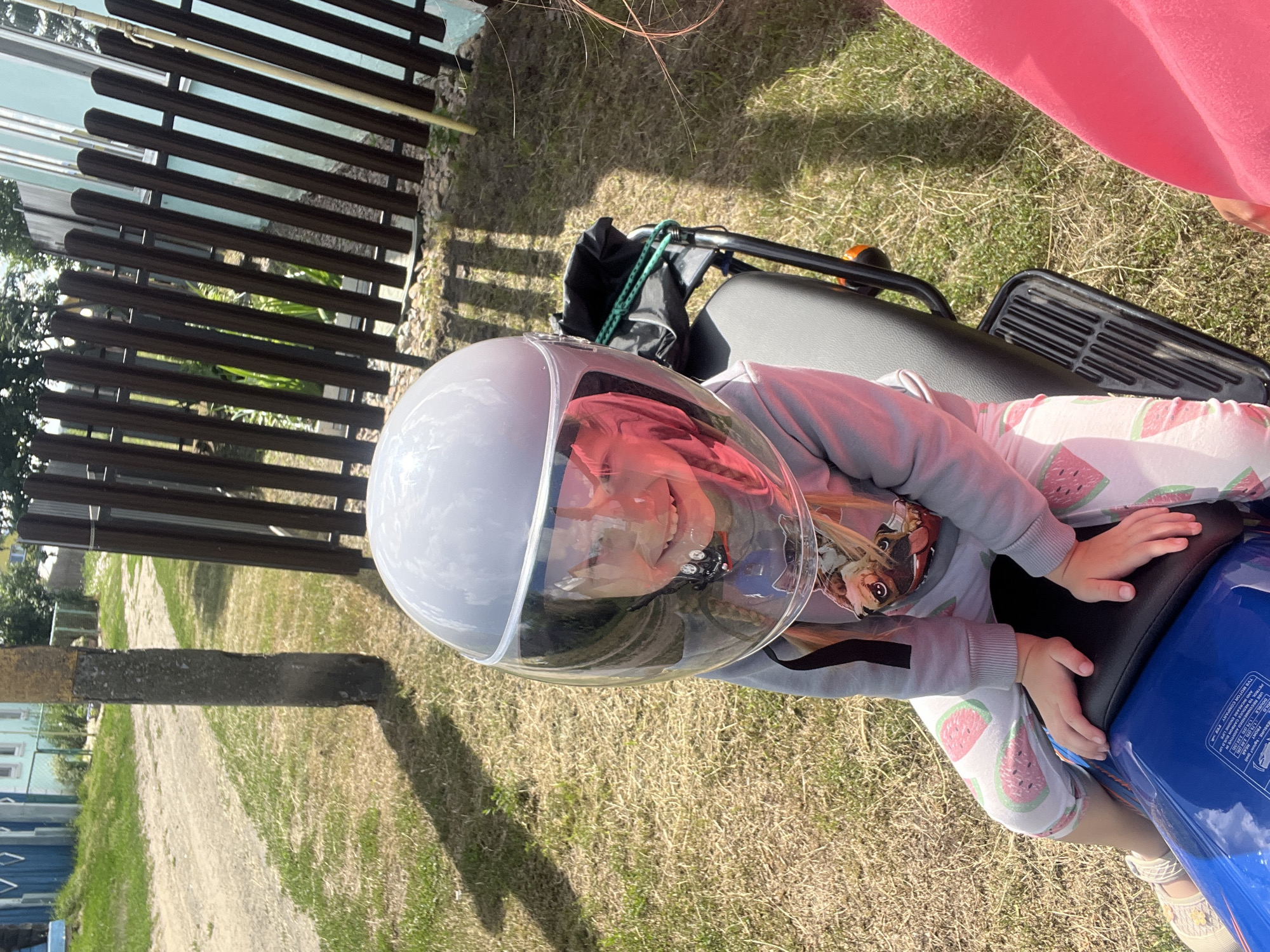 A young child, wearing a light gray hoodie with a graphic design on the front and white leggings with a watermelon pattern, is seated on a blue motorcycle. The child has a clear helmet on, and their face is visible inside. They appear to be smiling. Their hands are resting on the motorcycle seat.
The motorcycle is parked on a patch of dry, yellowed grass. In the background, a dark wooden fence is visible, and beyond that, some buildings and trees can be seen. The overall scene suggests an outdoor setting on a sunny day, possibly a backyard or a rural area. The time of day appears to be daytime, with bright sunlight casting shadows on the grass. There is some visible text on the blue part of the motorcycle, including "TVS MOTOR COMPANY" and tire pressure information.